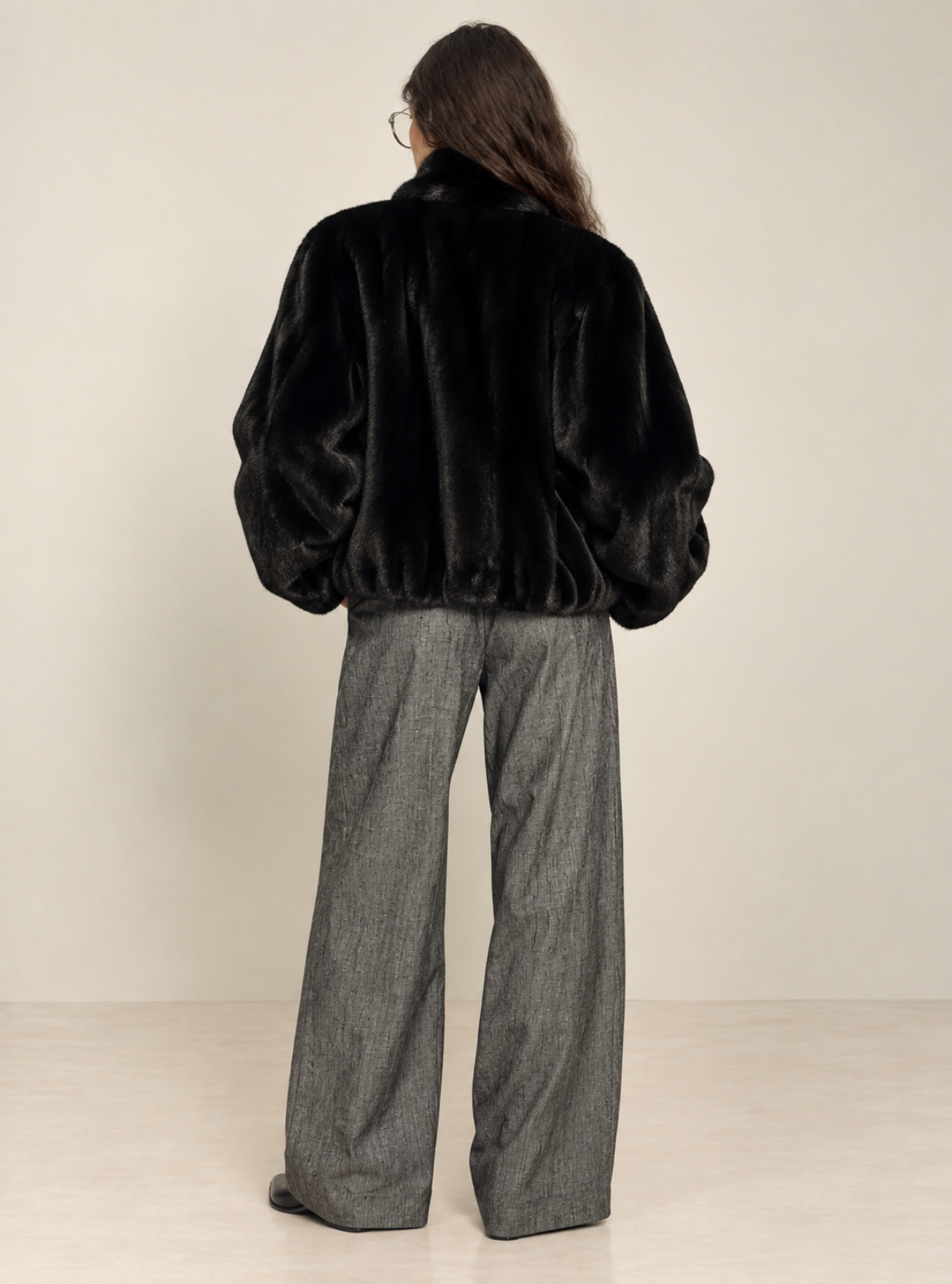 A model viewed from behind wearing a black faux fur cropped jacket and wide-leg gray trousers, standing against a plain beige studio background.