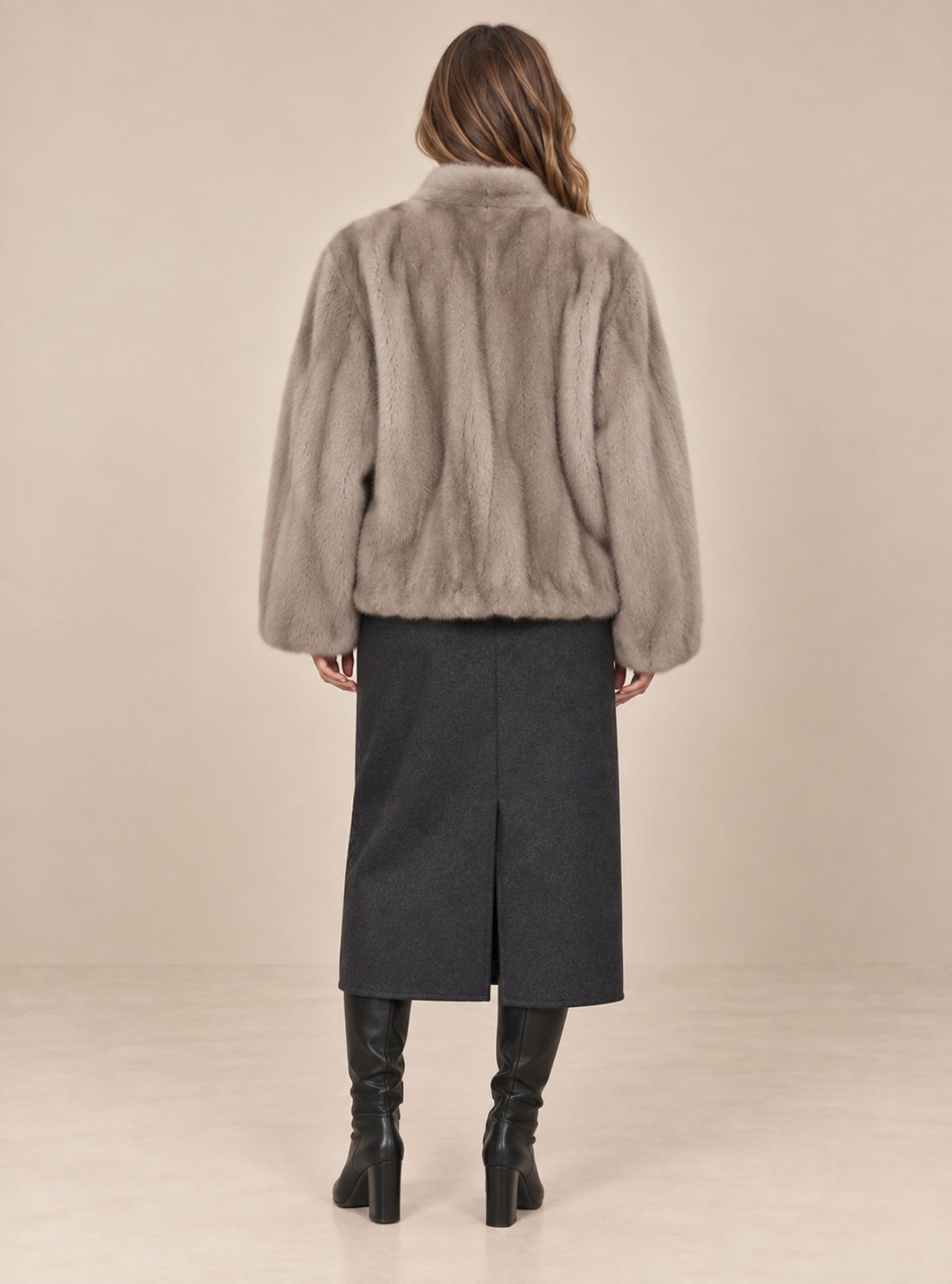 A woman shown from the back wearing a light taupe-gray fur jacket, a dark charcoal gray skirt, and black knee-high boots, set against a plain beige studio background.