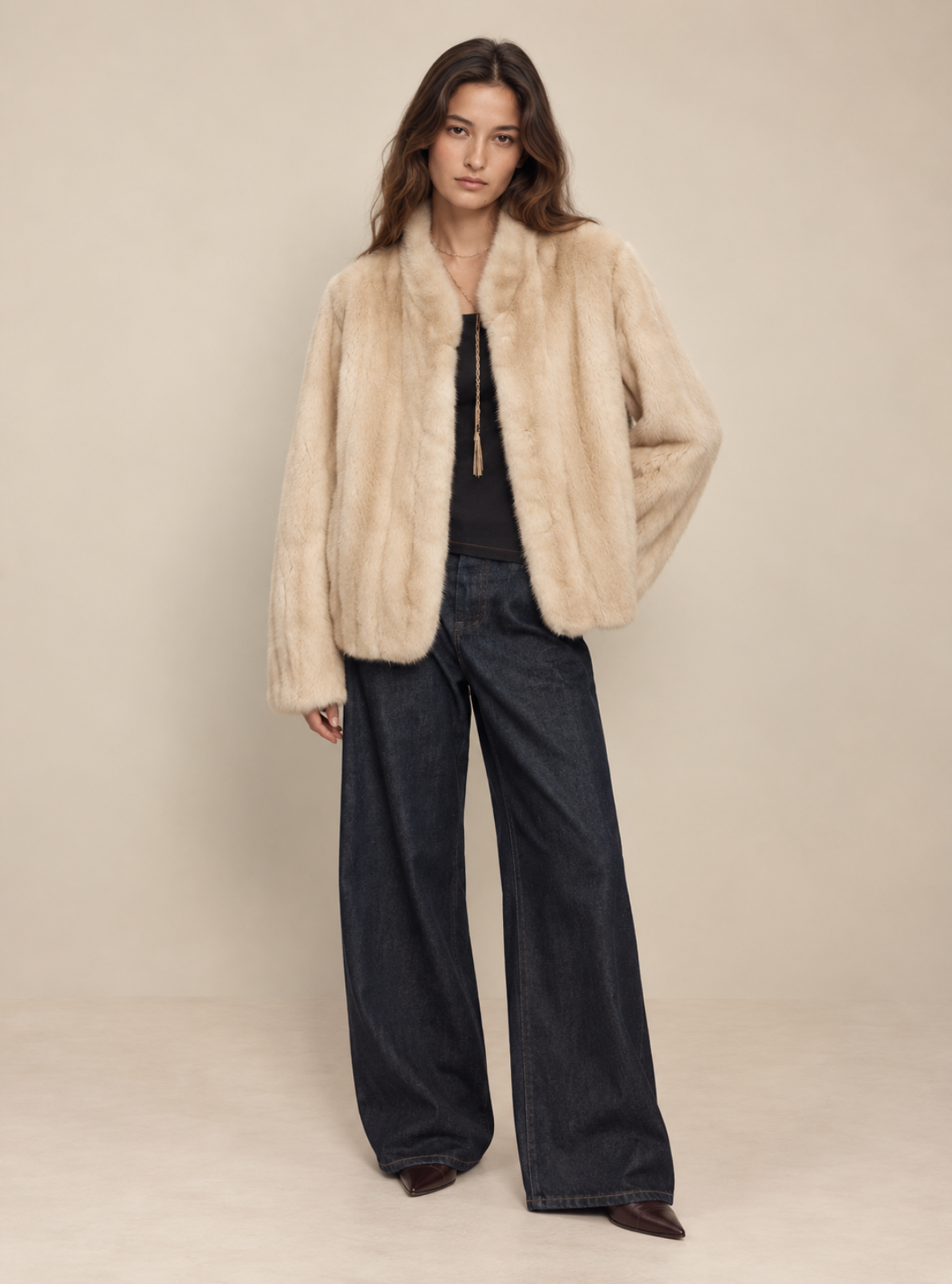 A model stands against a warm beige studio backdrop wearing a light beige faux-fur jacket over a black top and dark wide-leg jeans.