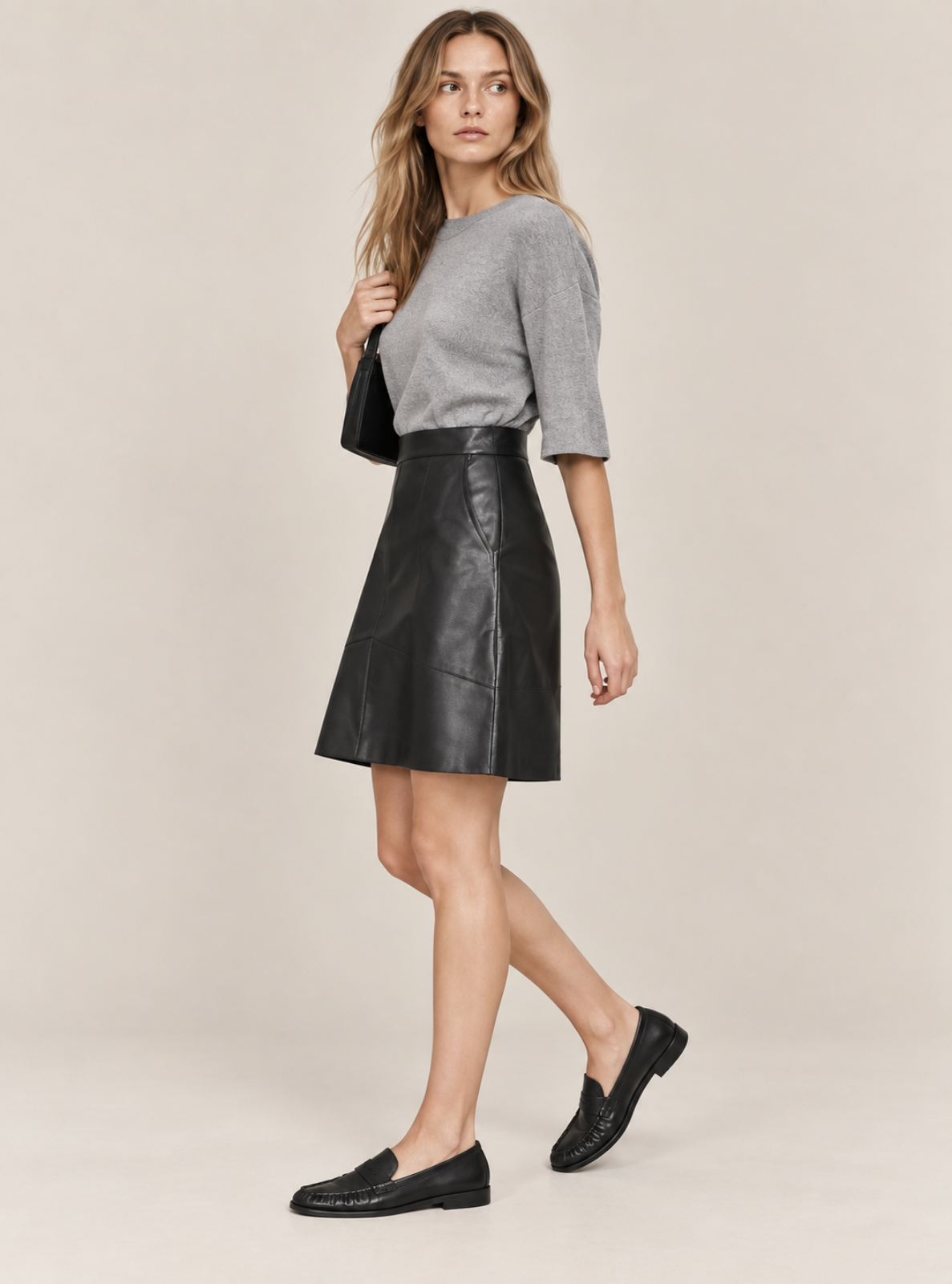 A woman modeling a black leather A-line mini skirt paired with a gray knit sweater, black loafers, and a small black shoulder bag, set against a plain beige studio background.
