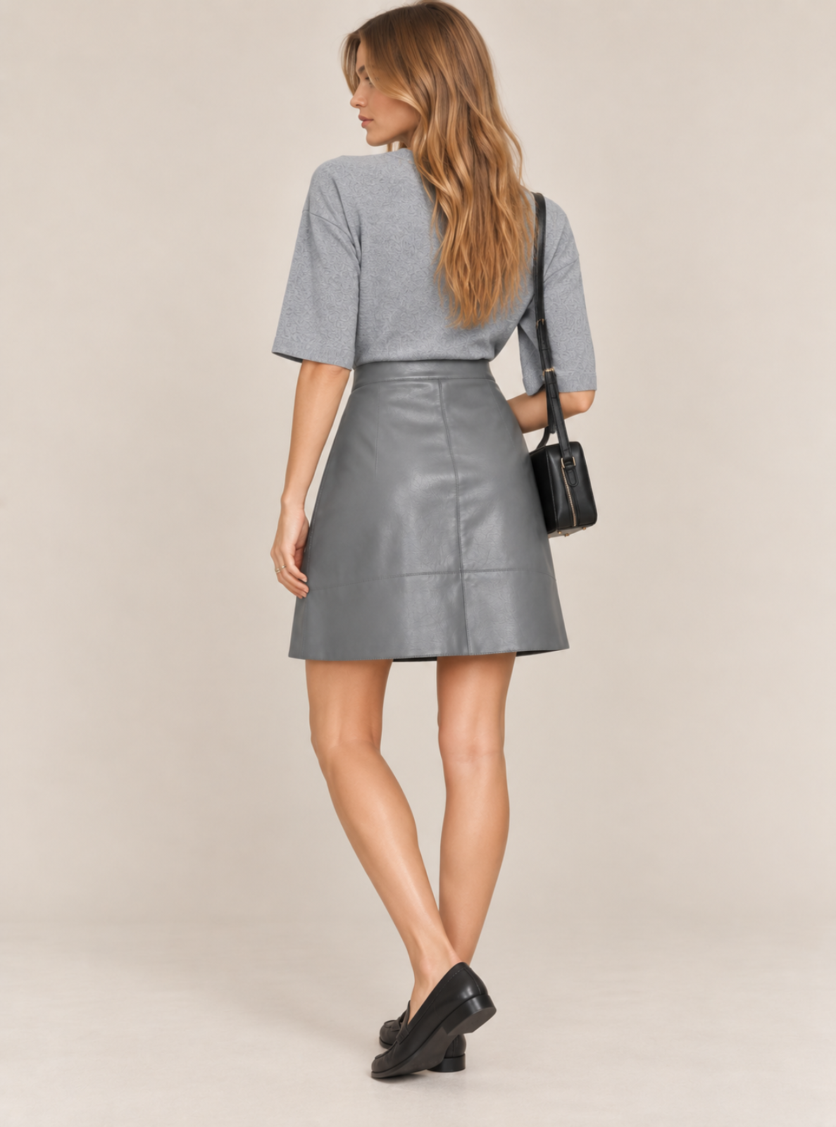 A woman is shown from the back wearing a gray short-sleeve knit top and a matching gray faux-leather A-line skirt, styled with a small black shoulder bag and black loafers, against a solid beige studio background.
