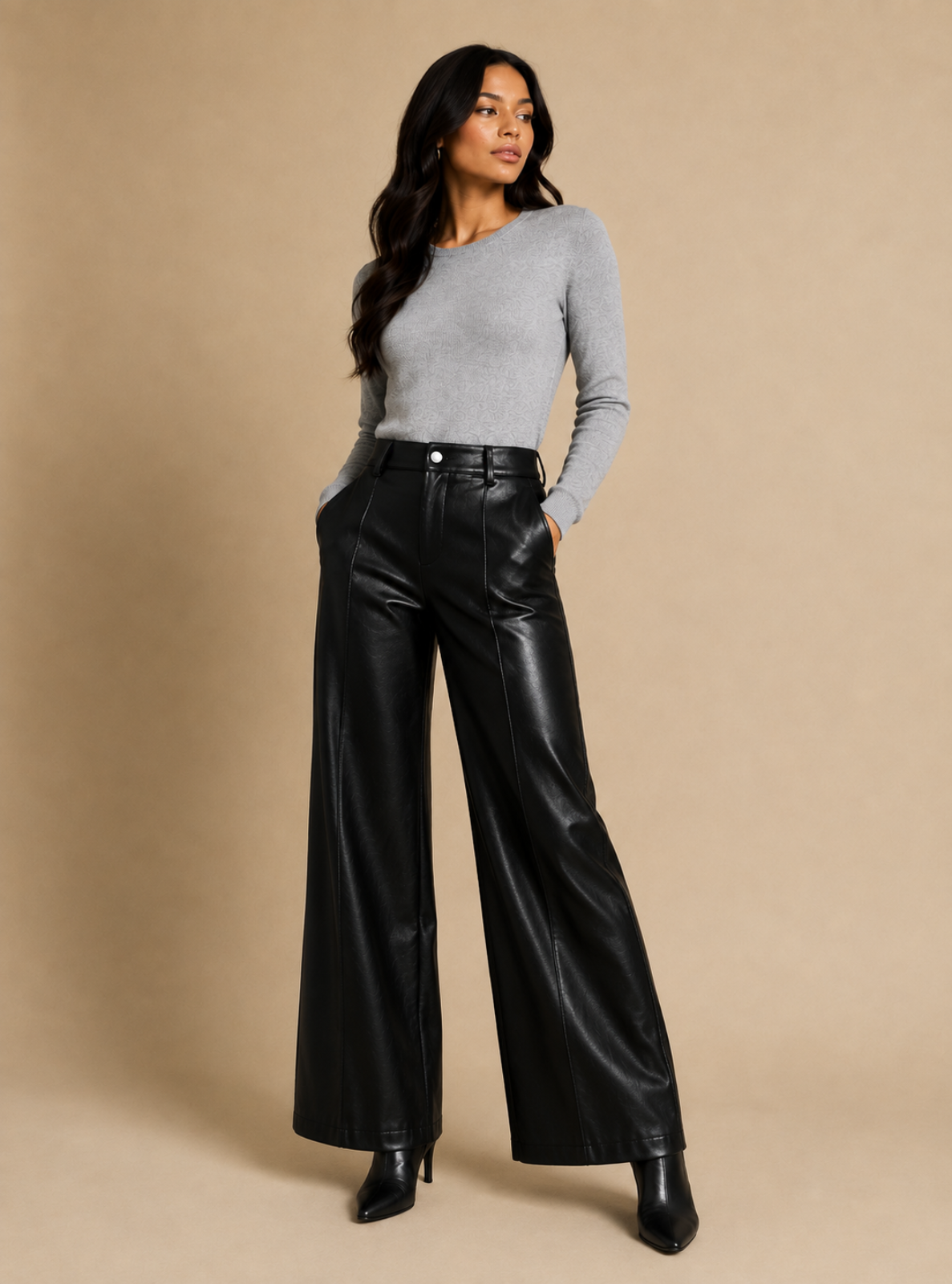 A woman modeling black wide-leg faux leather pants with a gray long-sleeve knit top and black ankle boots, standing against a solid beige studio backdrop.