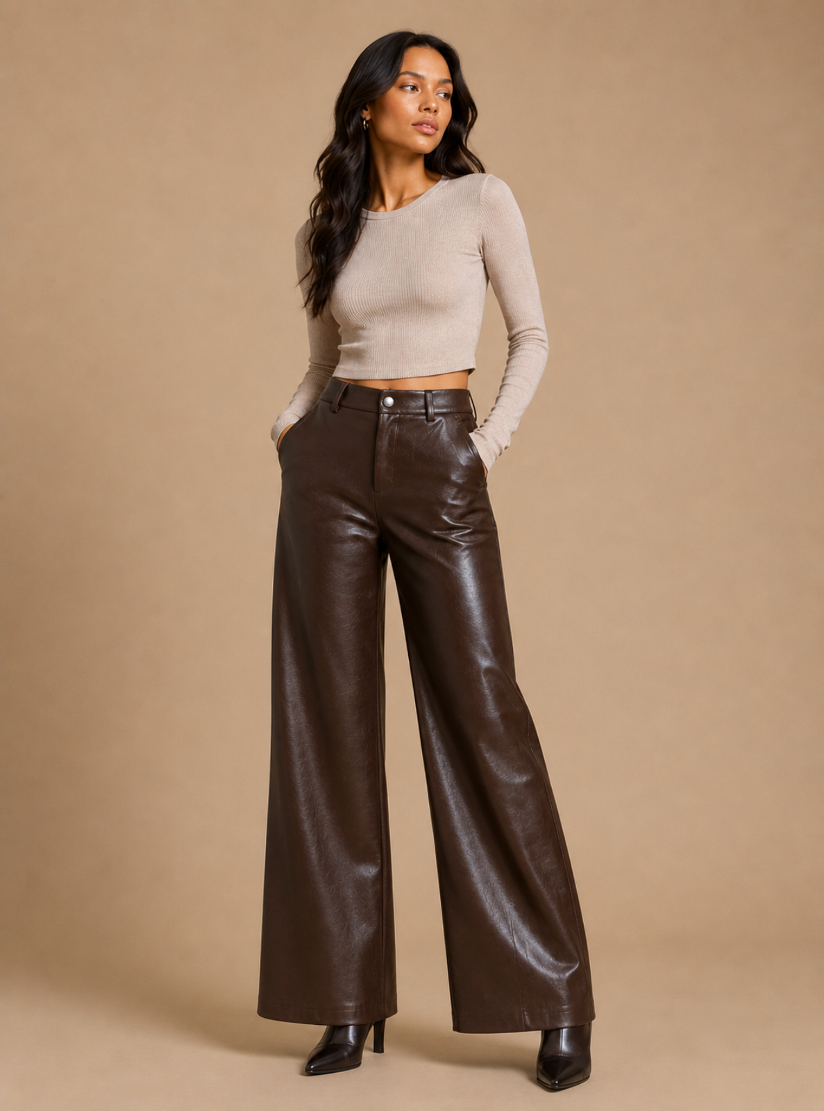 A woman modeling a beige long-sleeve crop top and dark brown wide-leg faux leather pants against a solid beige studio background.