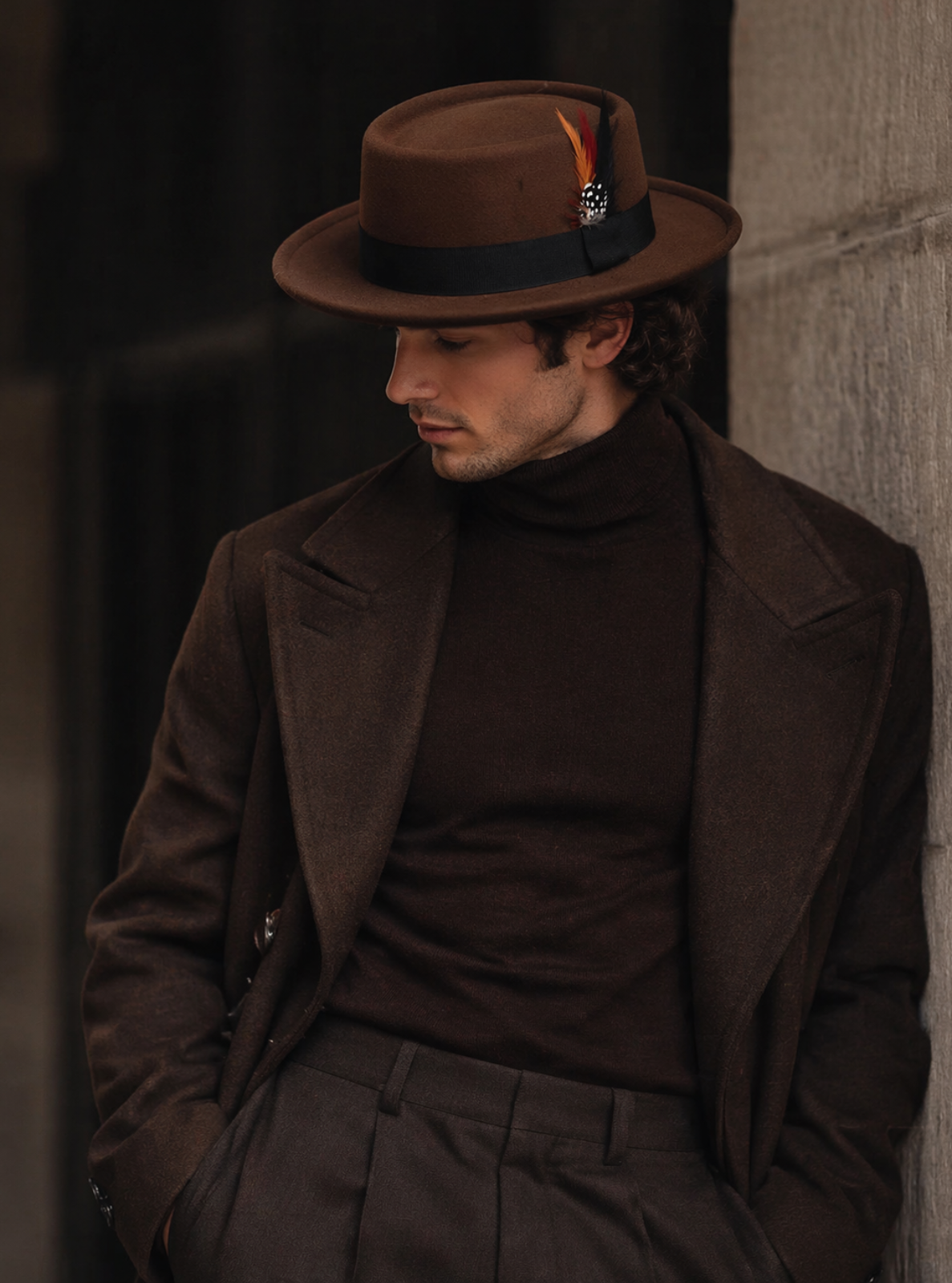 Man wearing a brown hat and coat leaning against a wall.