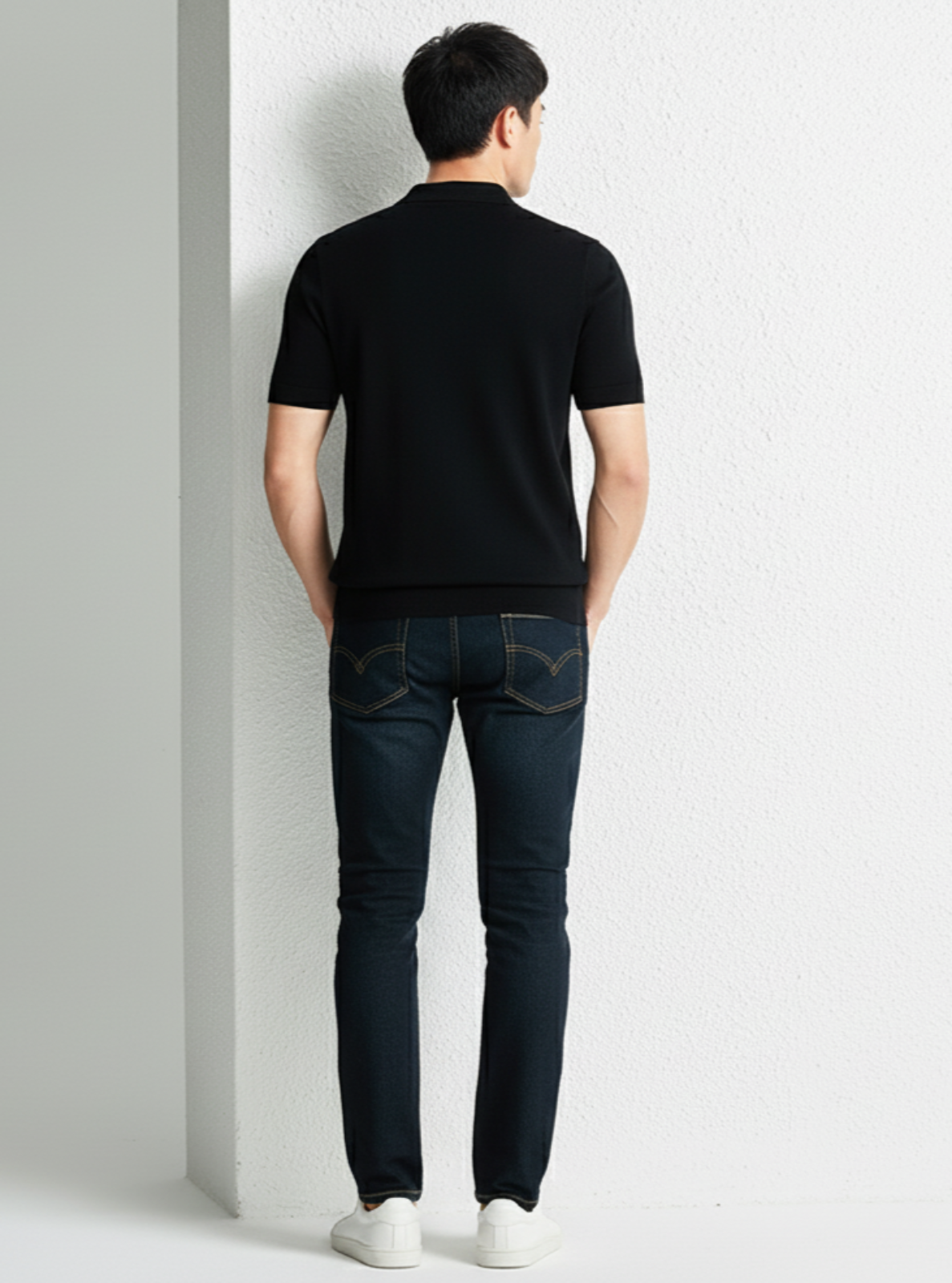 Person wearing a black t-shirt and dark jeans against a white wall