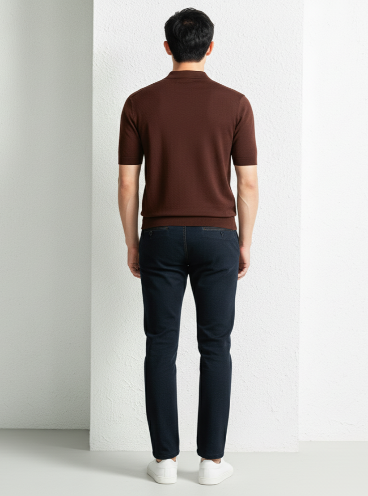 Man wearing a brown polo shirt and dark jeans against a white wall