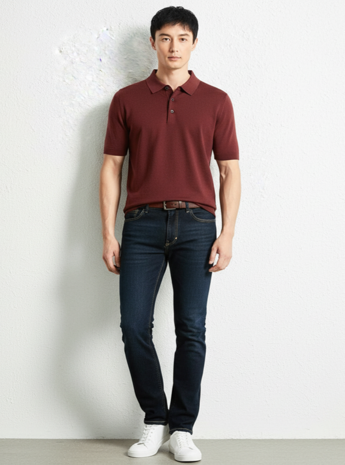 Man wearing a maroon polo shirt and dark jeans against a white wall