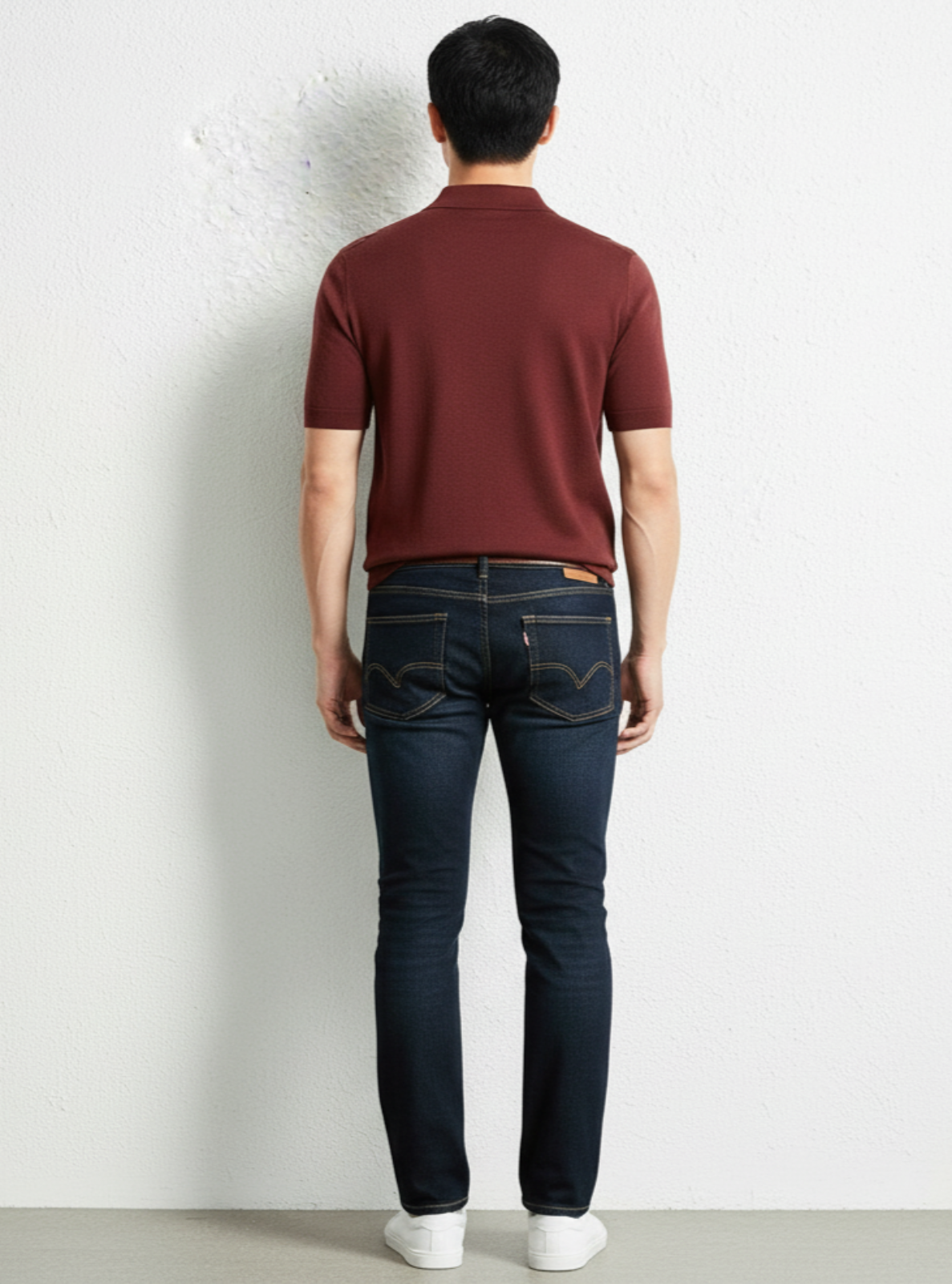 Man wearing a maroon polo shirt and dark jeans against a white wall.