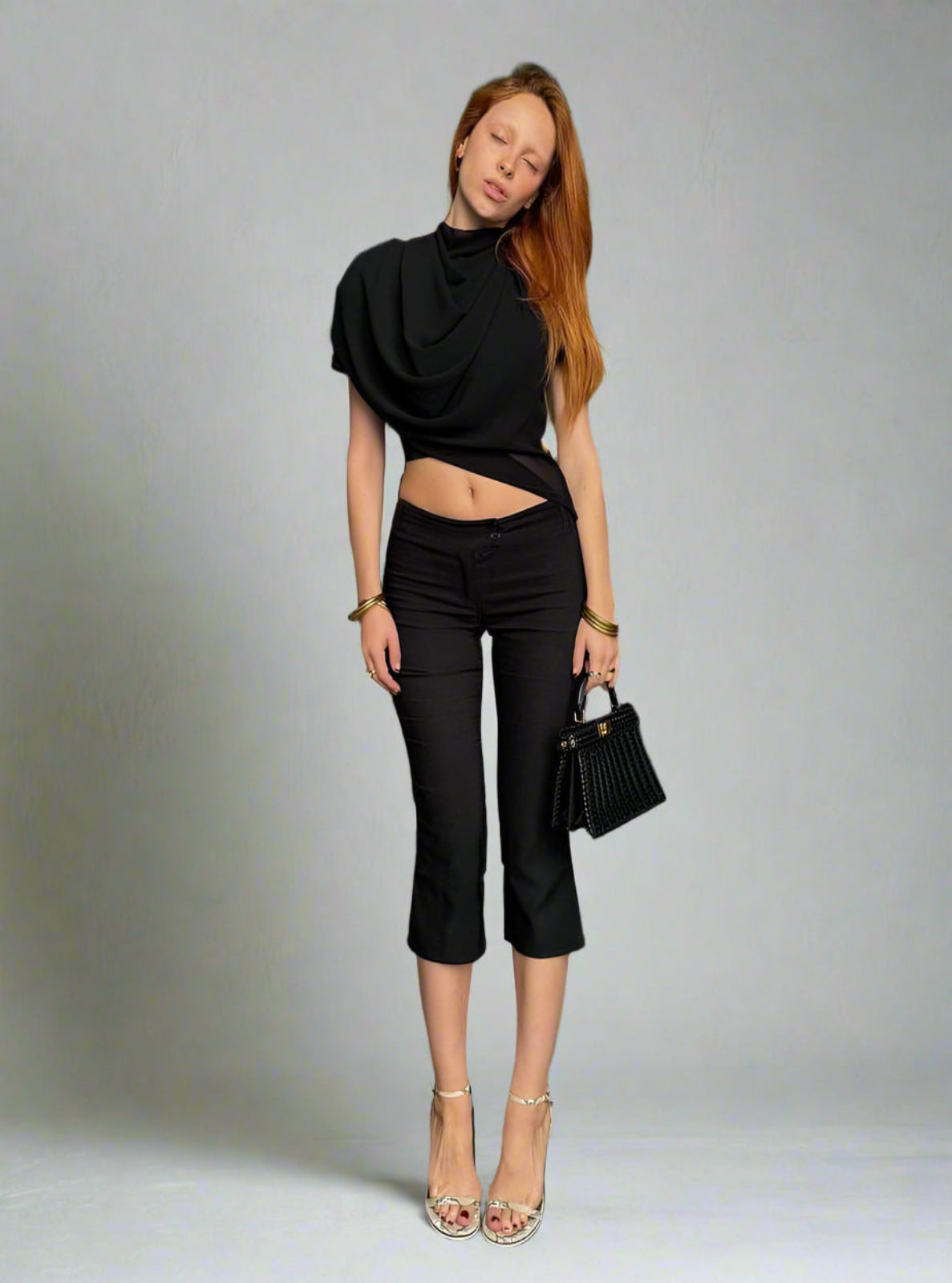 Woman wearing a black outfit with a draped top and high-waisted pants, holding a black handbag on a gray background.