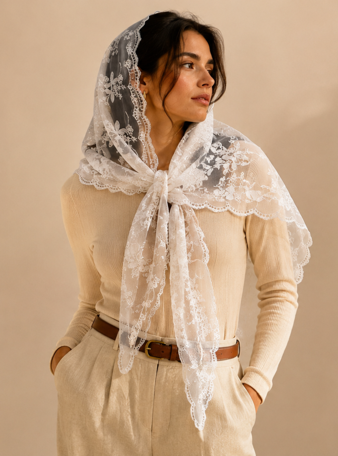 A woman wearing a cream-colored lace headscarf with a wide lace collar draped over her shoulders, styled with a beige sweater, cream trousers, and a brown belt against a warm beige background.