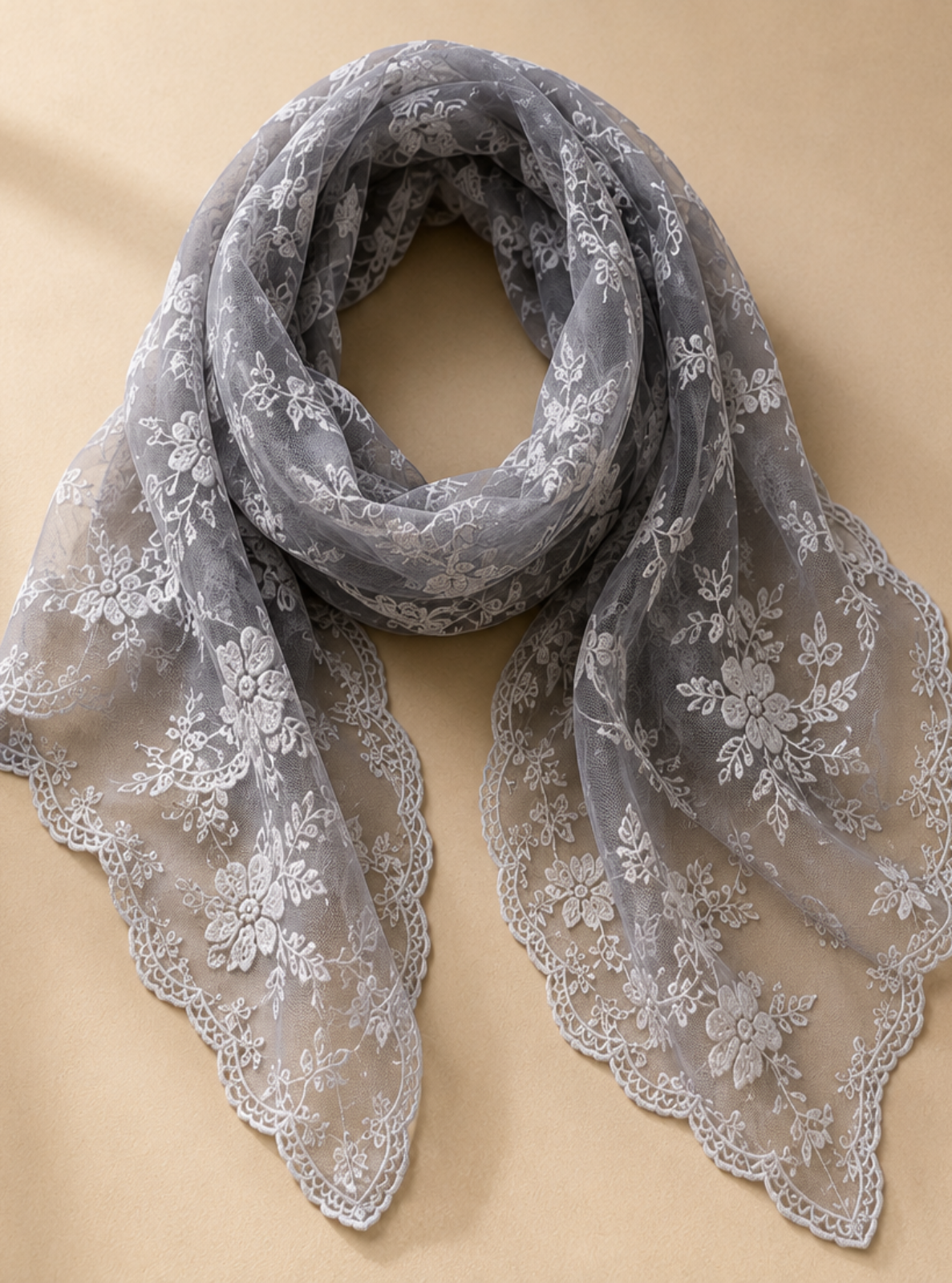 A gray sheer lace scarf with white floral embroidery and scalloped edges, draped on a warm beige background with soft window shadow overlays.
