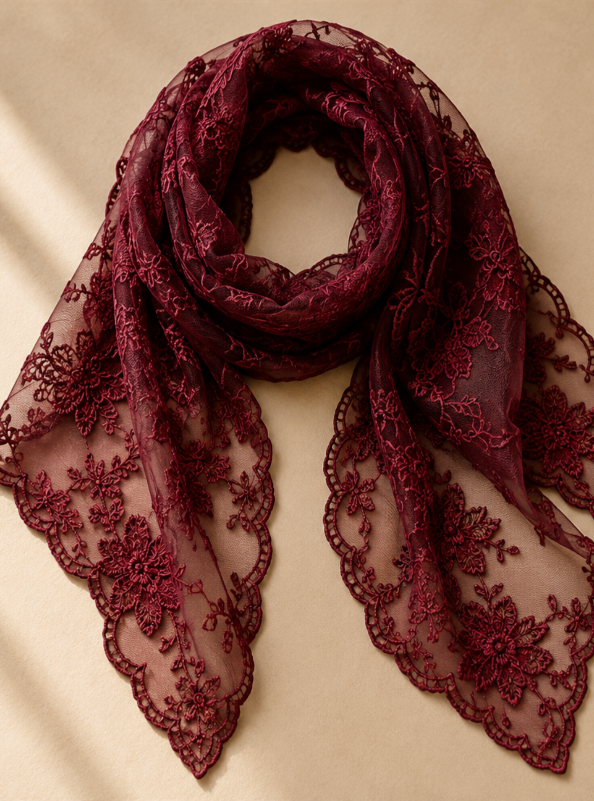 A burgundy lace scarf with floral embroidery, draped in a loop on a warm beige background with soft window shadow overlays.