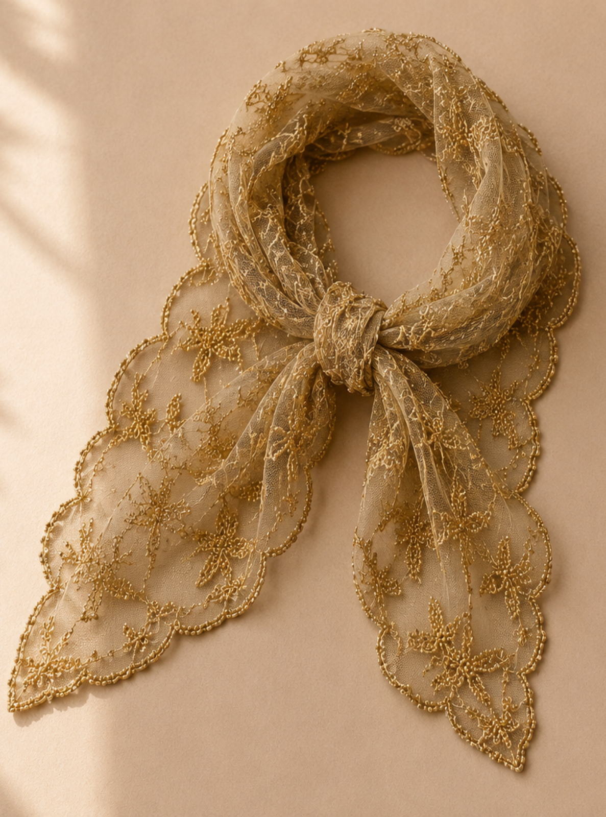A beige sheer lace scarf with embroidered floral details, tied in a bow and draped to show its scalloped edges, set against a warm beige background with soft leaf shadows.