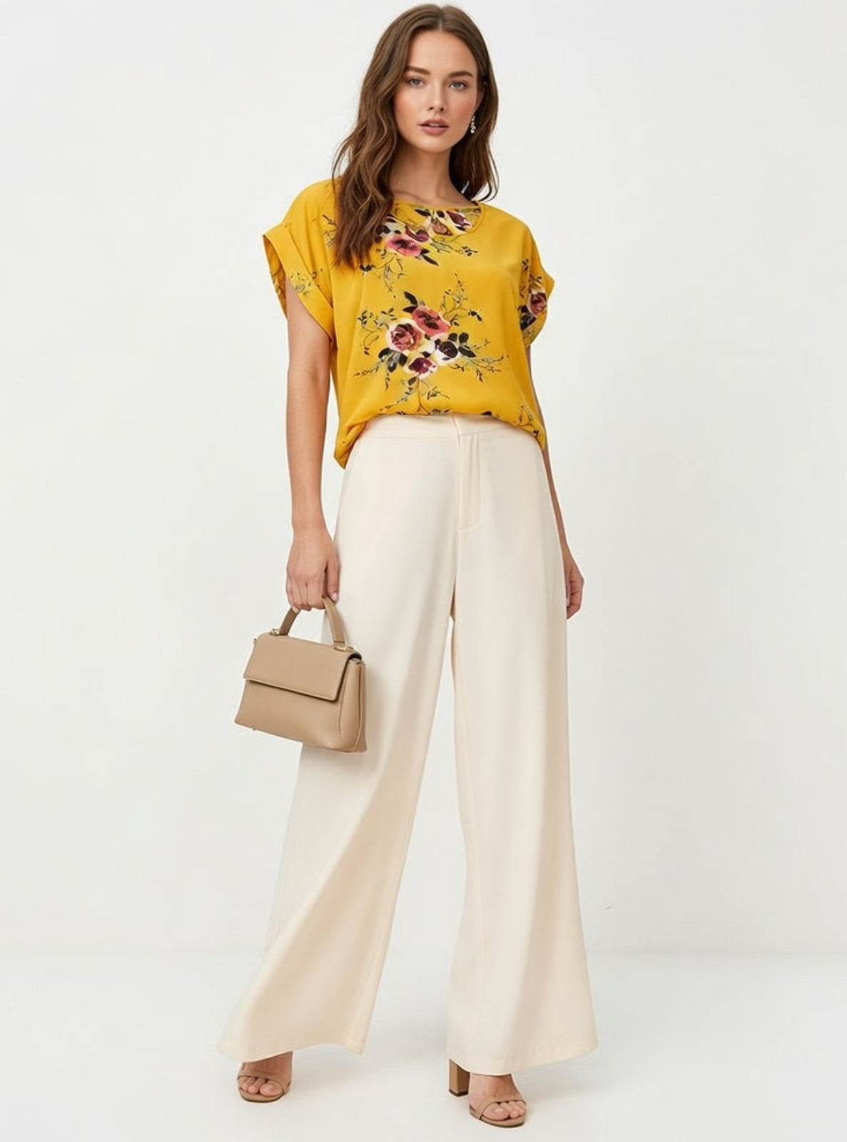 Woman wearing a yellow floral blouse and white pants, holding a beige handbag on a white background