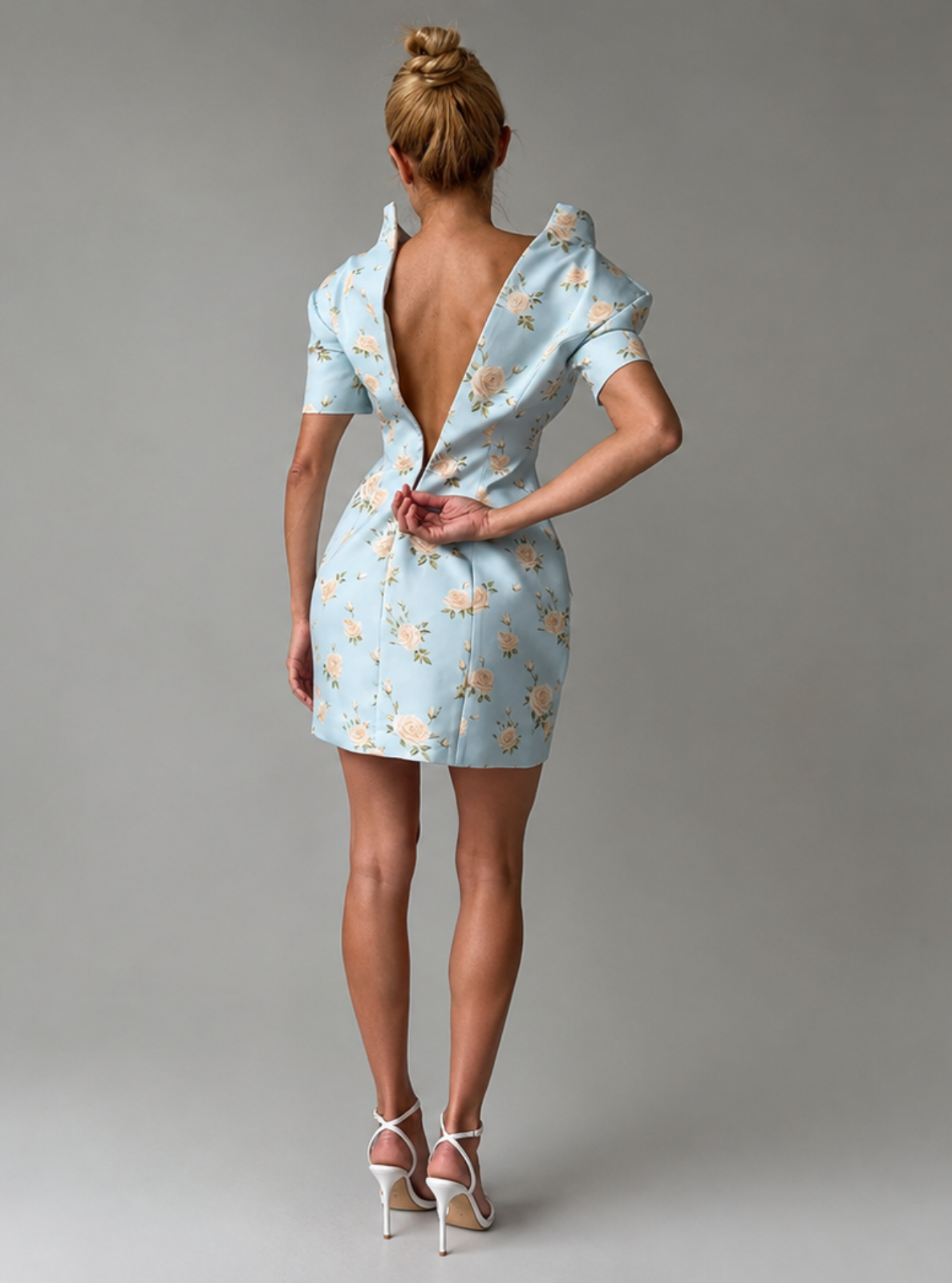 A woman stands with her back to the camera wearing a light blue short dress with a yellow floral pattern and a very large open back. She is wearing white high-heeled sandals against a neutral gray studio background.
