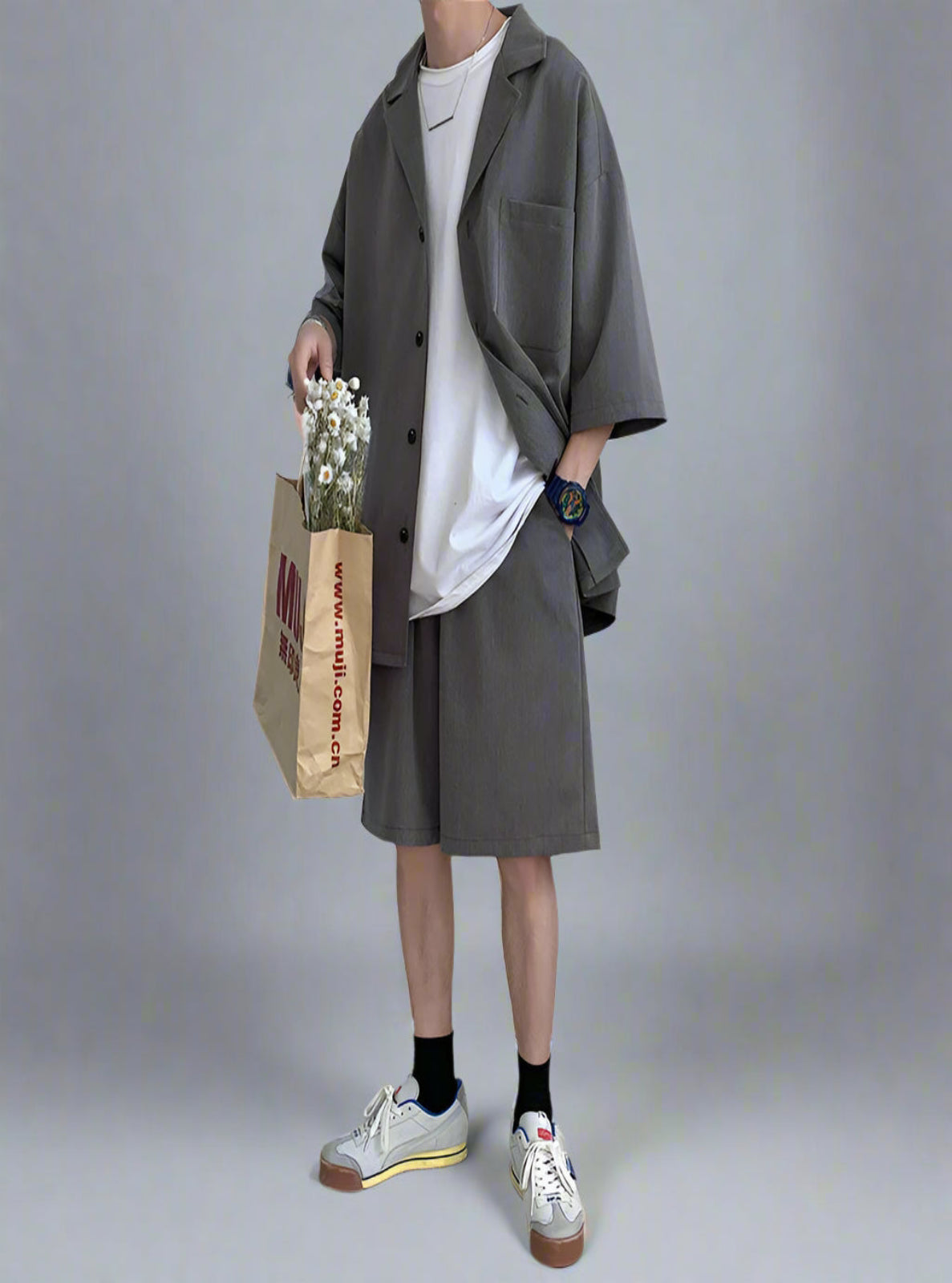 Person wearing a gray coat and white sneakers holding a paper bag with flowers against a white wall.