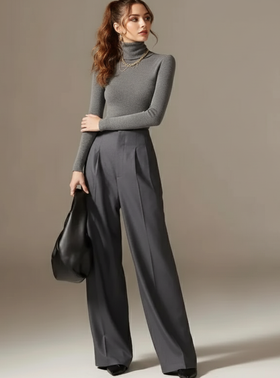 Woman in gray turtleneck and wide-leg pants holding a black handbag against a neutral background