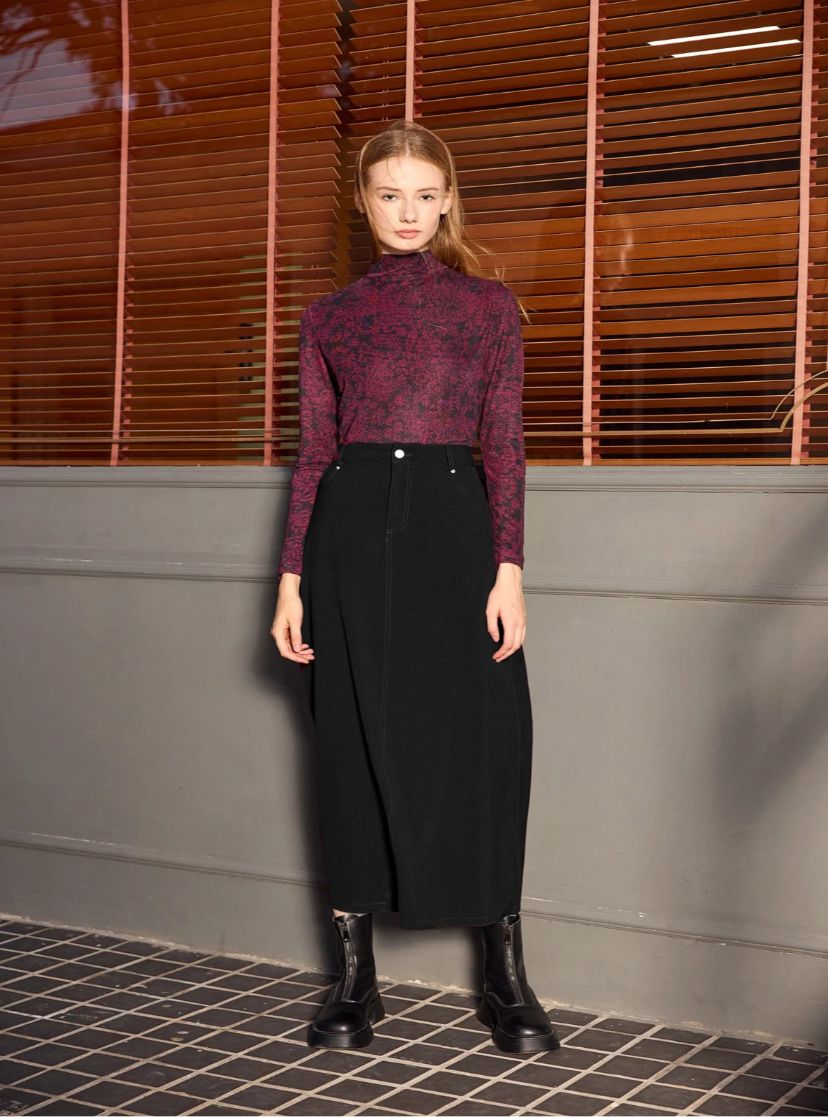 Woman wearing a patterned top and black skirt standing against a wall with wooden slats.