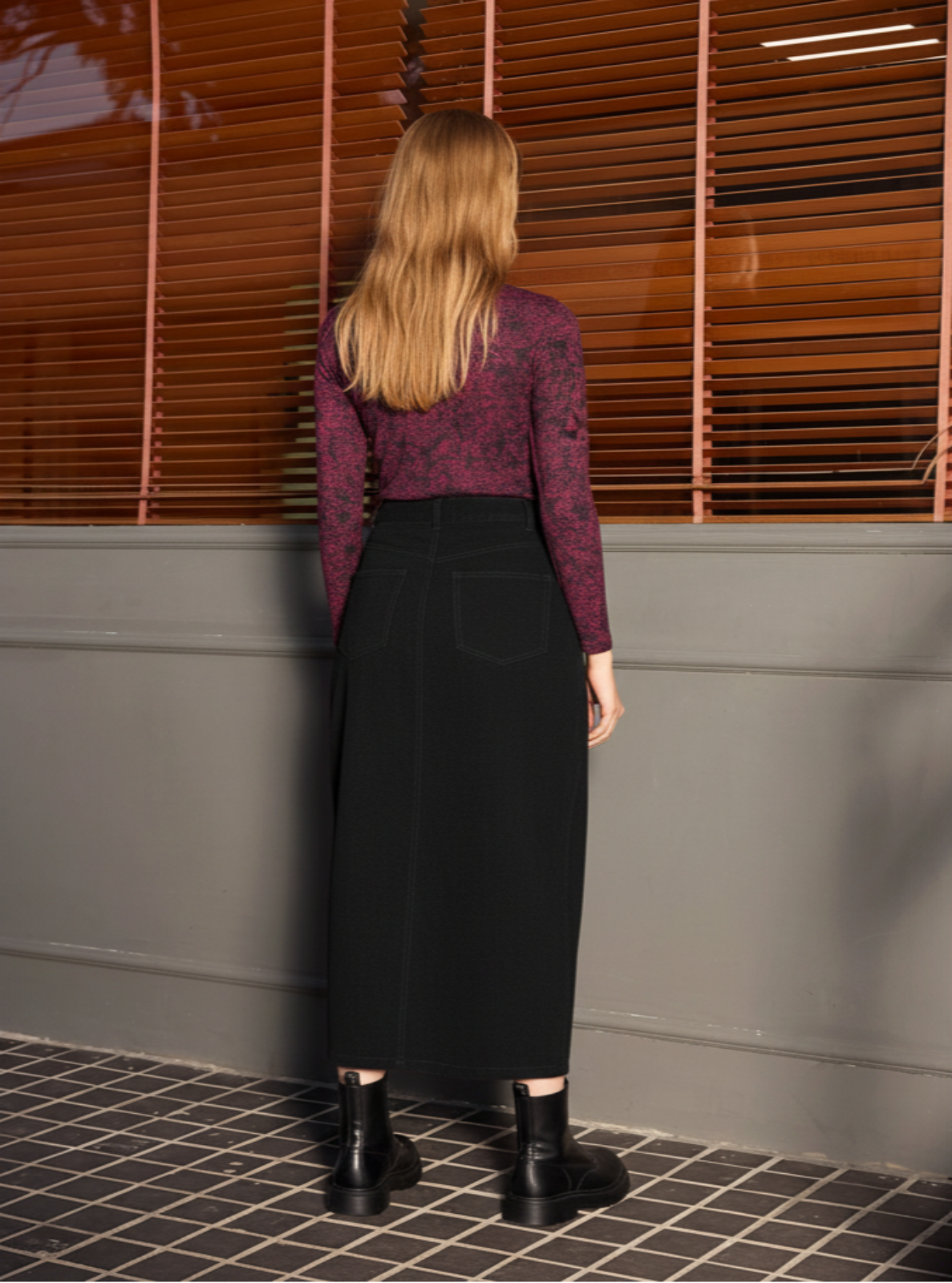 Woman wearing a patterned top and black skirt standing against a wall with horizontal slats.