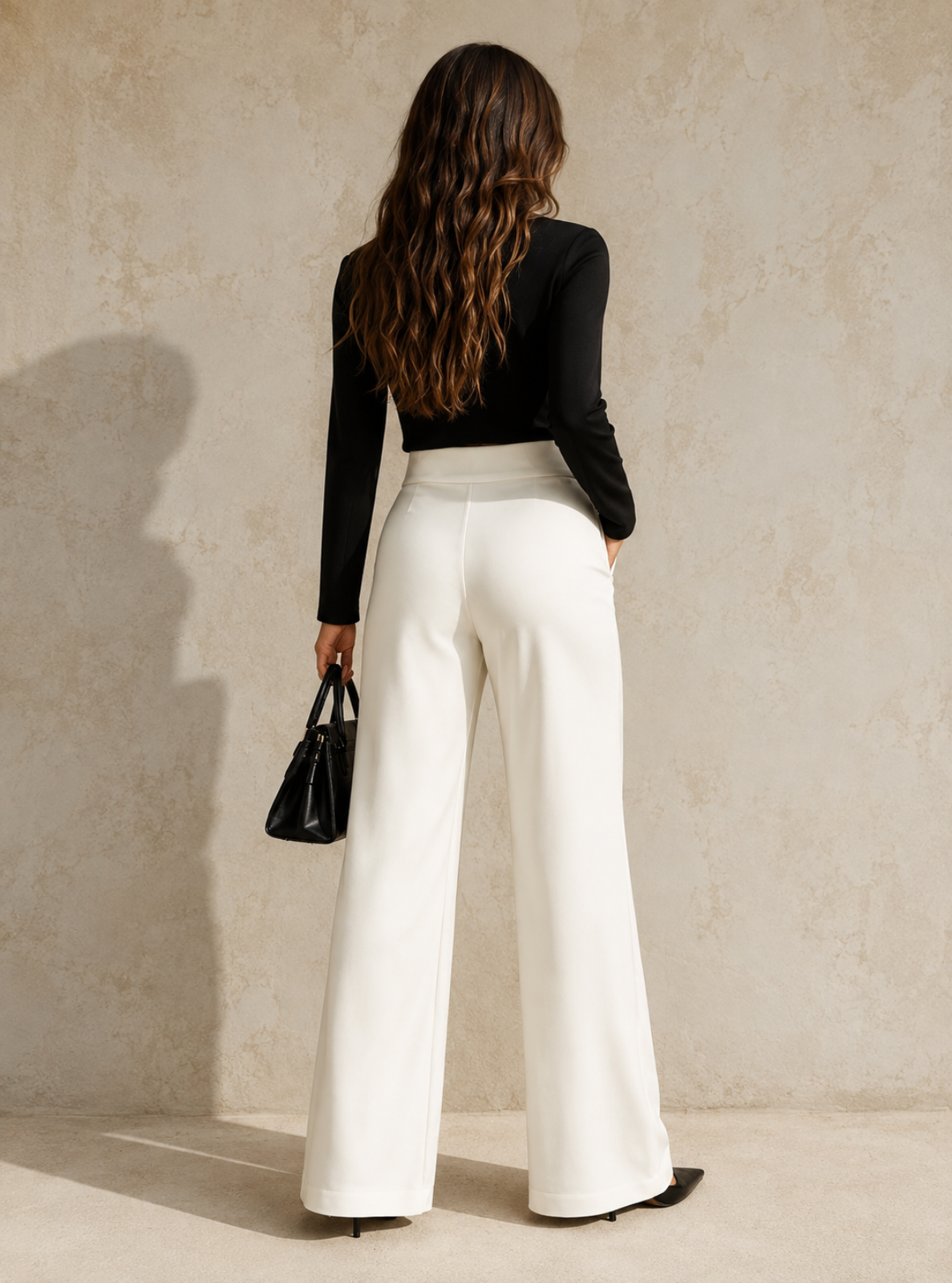 A woman shown from behind wearing high-waisted, wide-leg white trousers and a black long-sleeve top, holding a small black handbag against a textured beige wall.