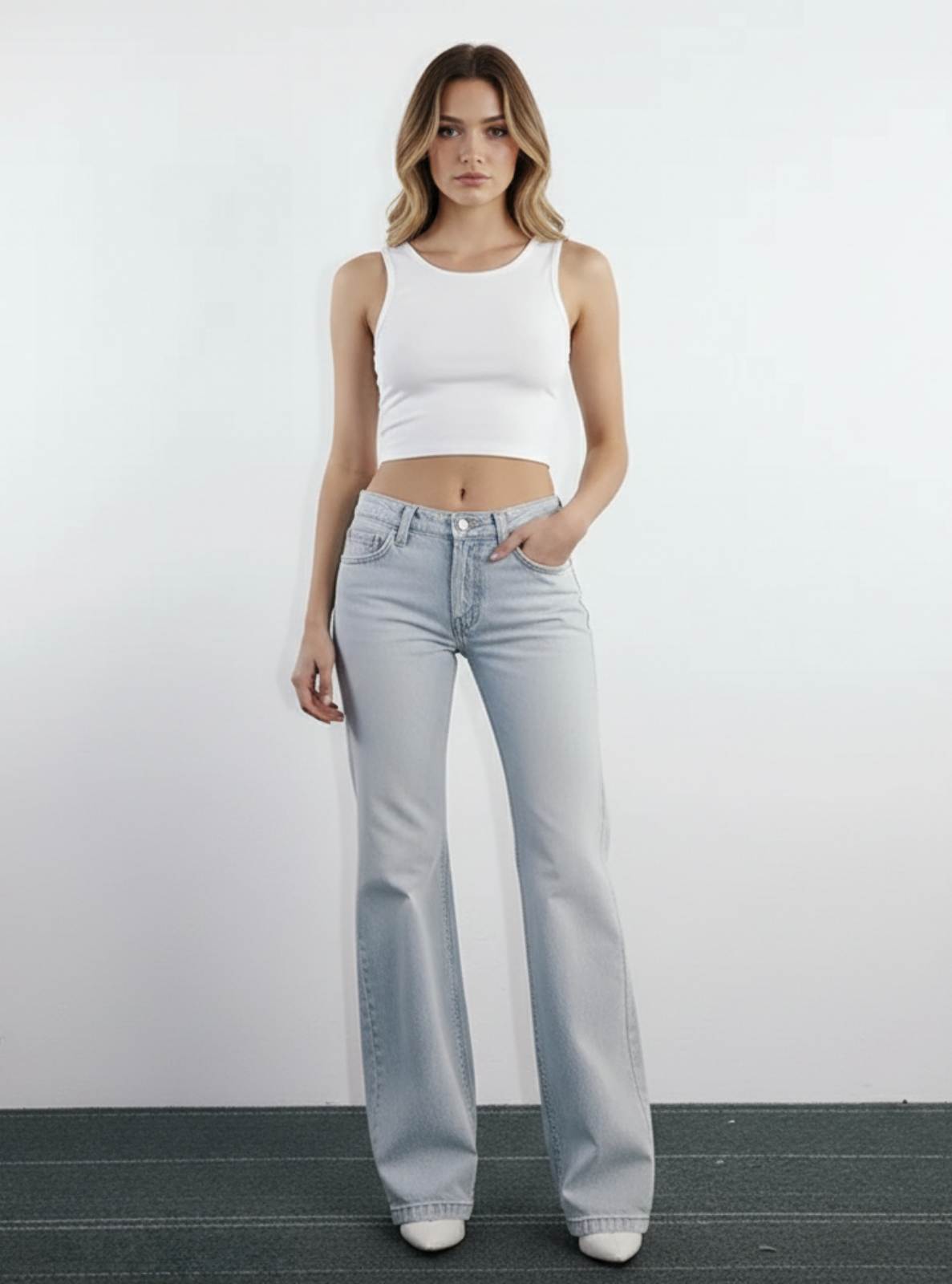 Woman wearing light blue jeans and a white tank top against a plain background