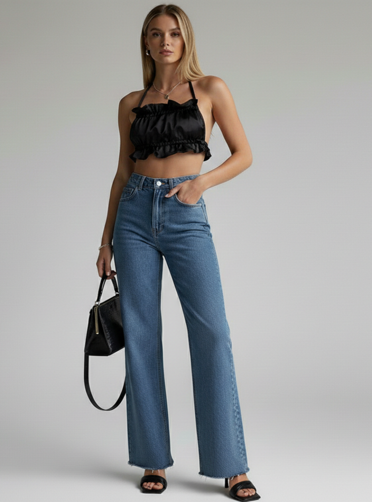 Woman wearing a black top and blue jeans holding a black handbag on a gray background