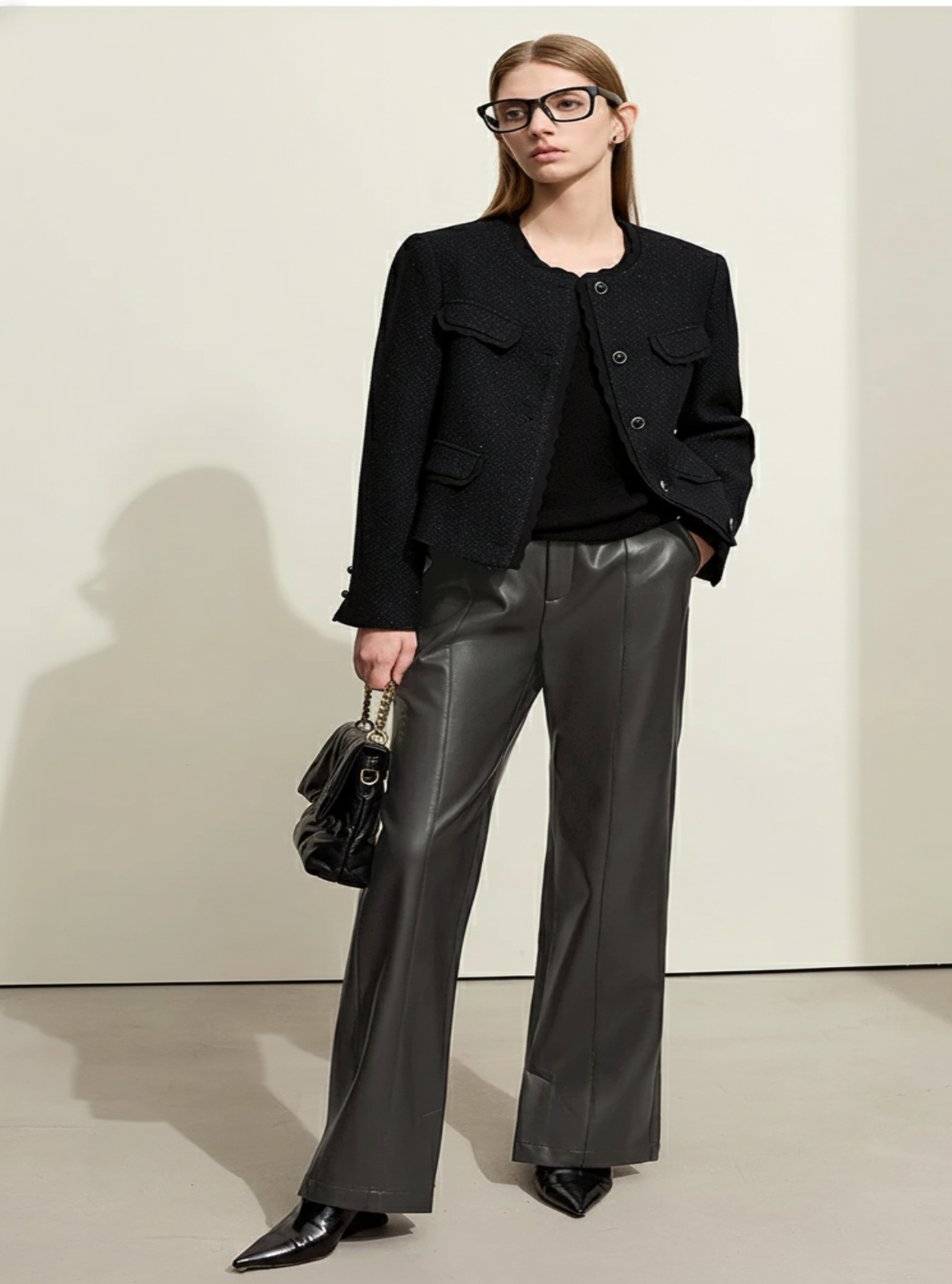Woman wearing a black jacket and leather pants holding a black handbag against a white background