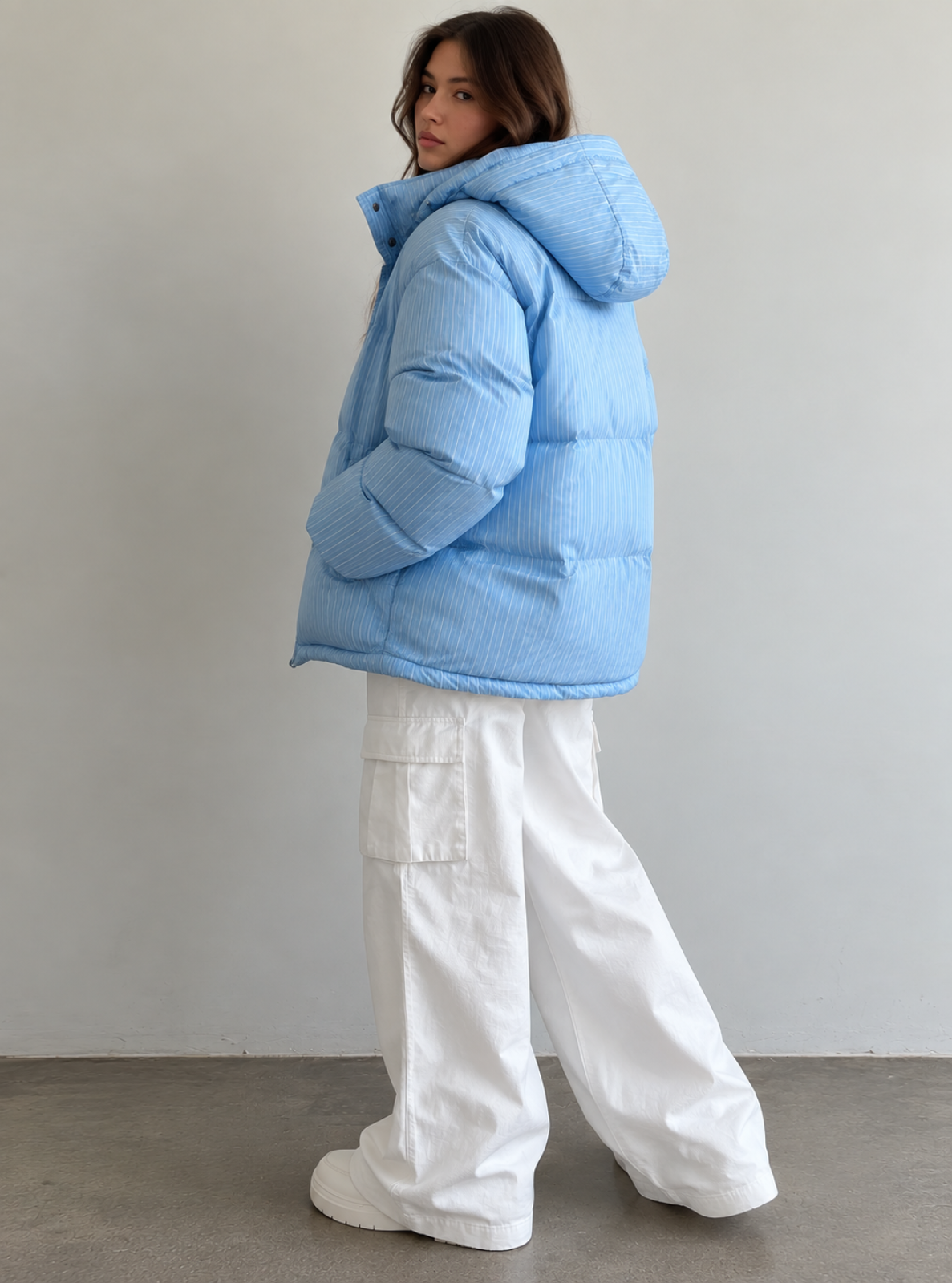 A woman wearing a light blue puffer jacket with a hood and white wide-leg cargo pants, standing against a plain light gray wall.