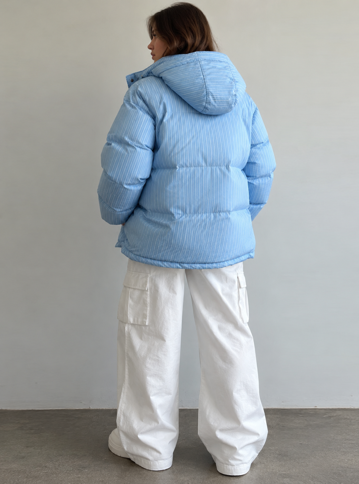 A person shown from the back wearing a light blue puffer jacket with a hood and white wide-leg cargo pants, standing against a plain light gray wall.