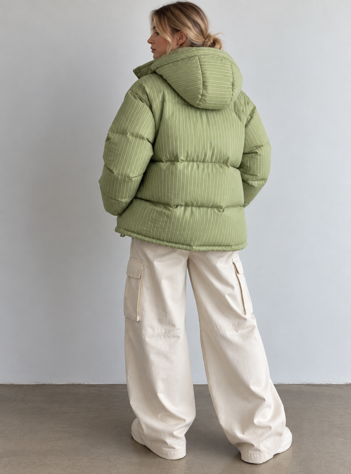 A woman shown from the back wearing a light sage green puffer jacket with a ribbed texture and cream wide-leg cargo pants, standing against a plain light gray wall.