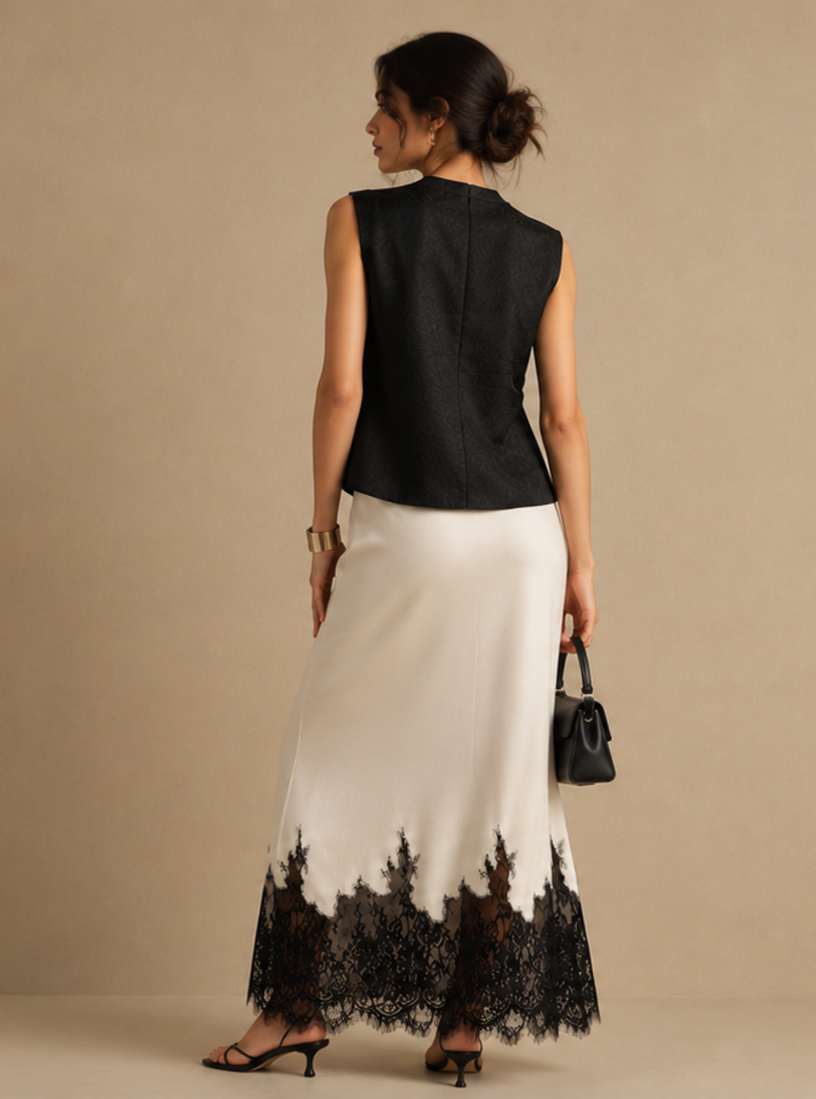 A woman stands with her back to the camera wearing a sleeveless black top and a long white skirt with a black lace hem, holding a small black handbag against a plain beige studio background.