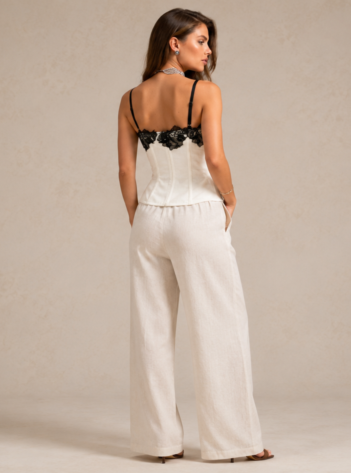 A woman stands with her back to the camera wearing a white wide-leg pants jumpsuit with a black lace corset-style top, set against a neutral beige studio backdrop.