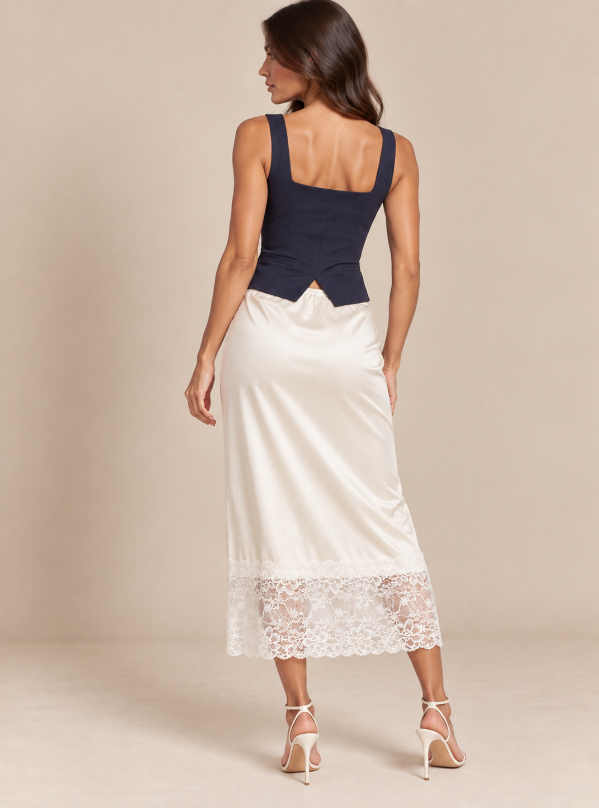 A woman shown from the back wearing a sleeveless midi dress with a dark navy blue structured bodice and a flowing ivory satin skirt featuring a lace hem, paired with white high-heeled sandals, set against a solid beige studio background.