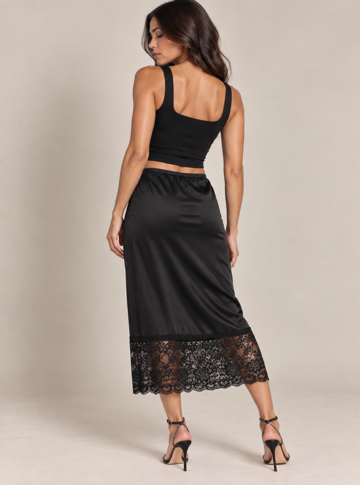 A woman modeling a black two-piece outfit consisting of a sleeveless crop top and a satin midi skirt with a lace hem, shown from the back against a neutral beige studio background.