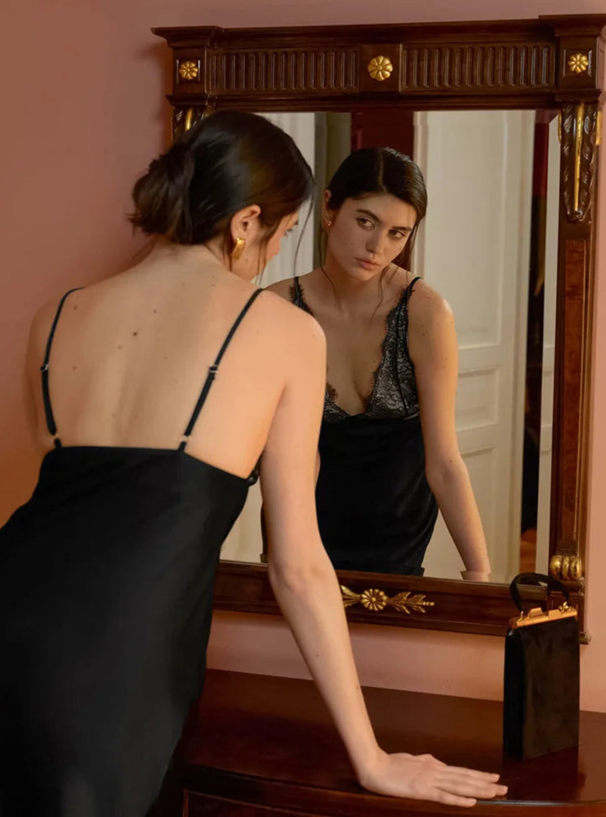 Woman in a black dress looking at herself in a large mirror.