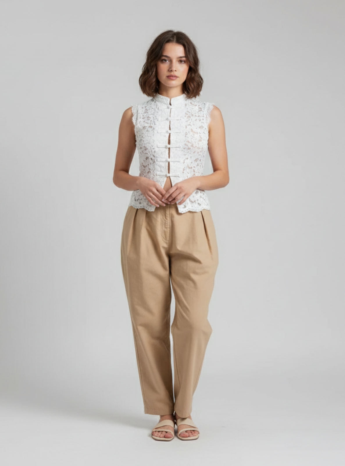 Woman wearing a white sleeveless top with lace details and beige pants on a light gray background