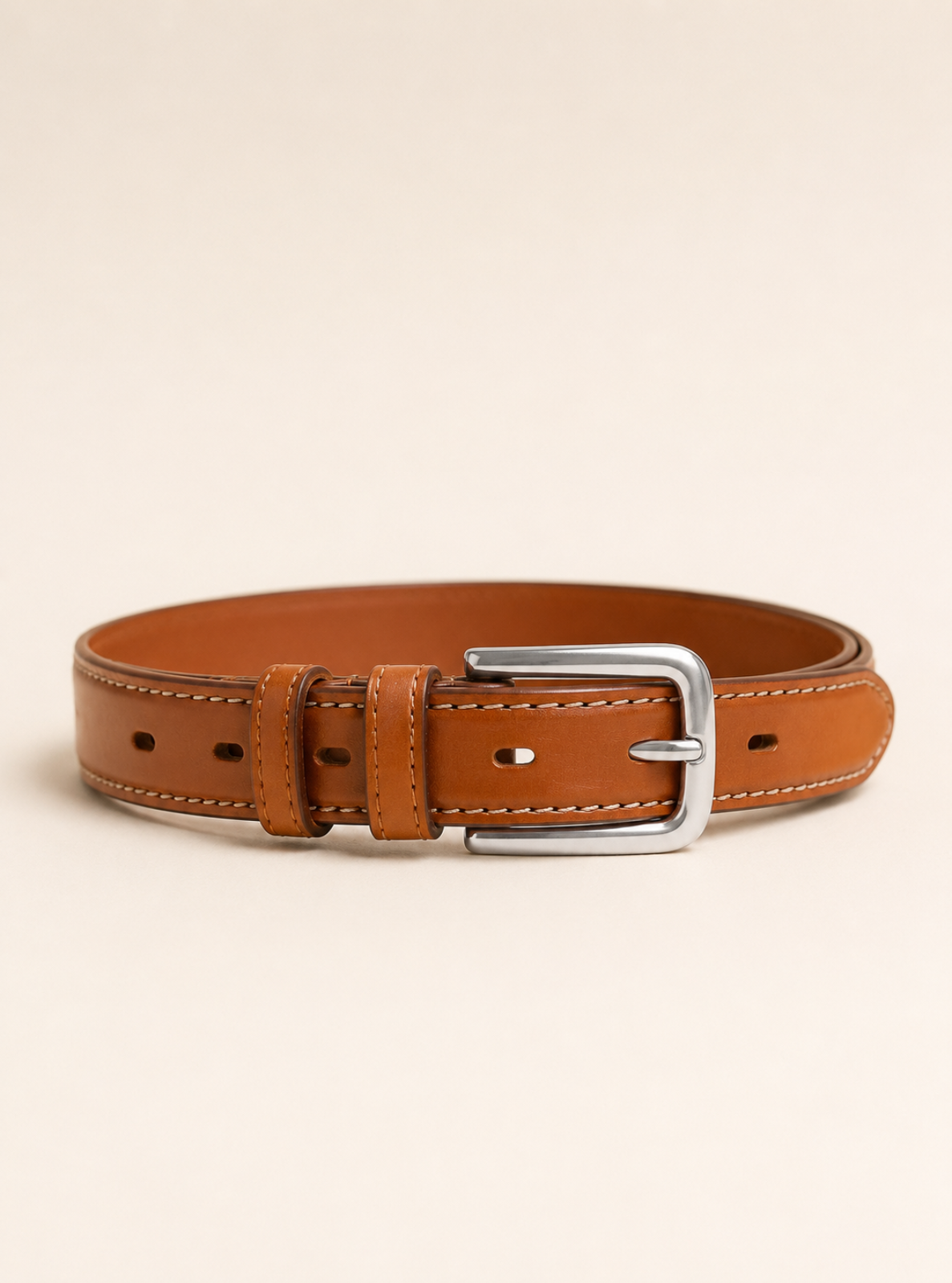 A tan leather belt with a silver metal buckle, neatly coiled and presented on a solid light beige background.