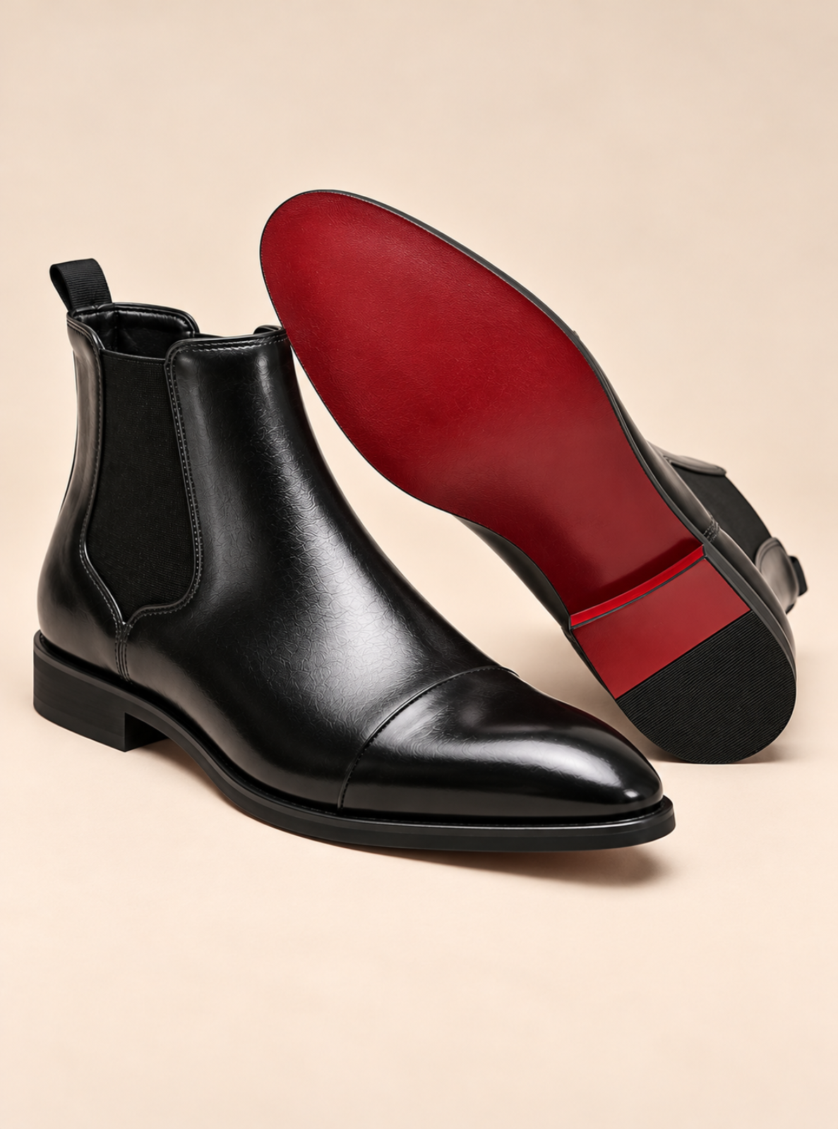 A pair of black leather Chelsea boots with elastic side panels, shown on a warm beige background. One boot is standing upright to show the black cap toe and low block heel, while the other is tilted on its side to reveal a contrasting red outsole.