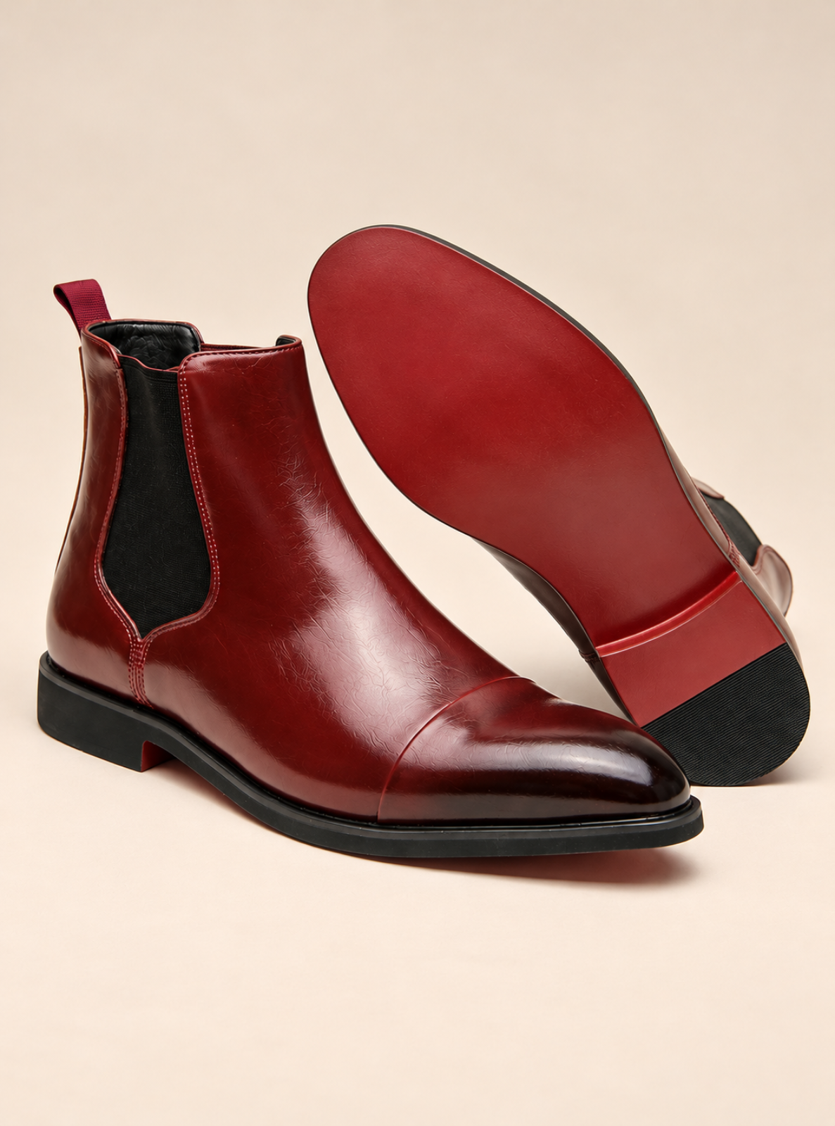 A pair of glossy dark red leather Chelsea boots, with one boot standing upright to show the side and the other lying on its side to display the red outsole and black heel.