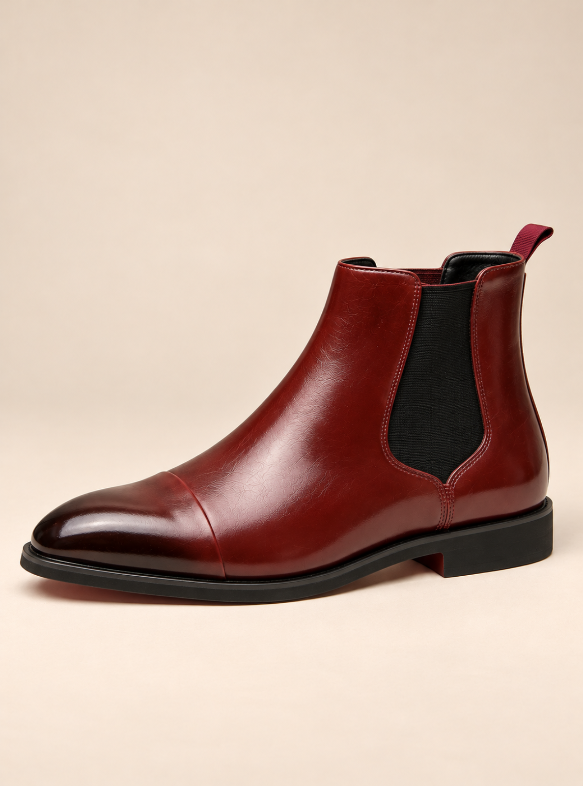 A single men's Chelsea-style ankle boot in glossy burgundy leather with black elastic side panels and a black sole, photographed on a plain warm beige studio background.