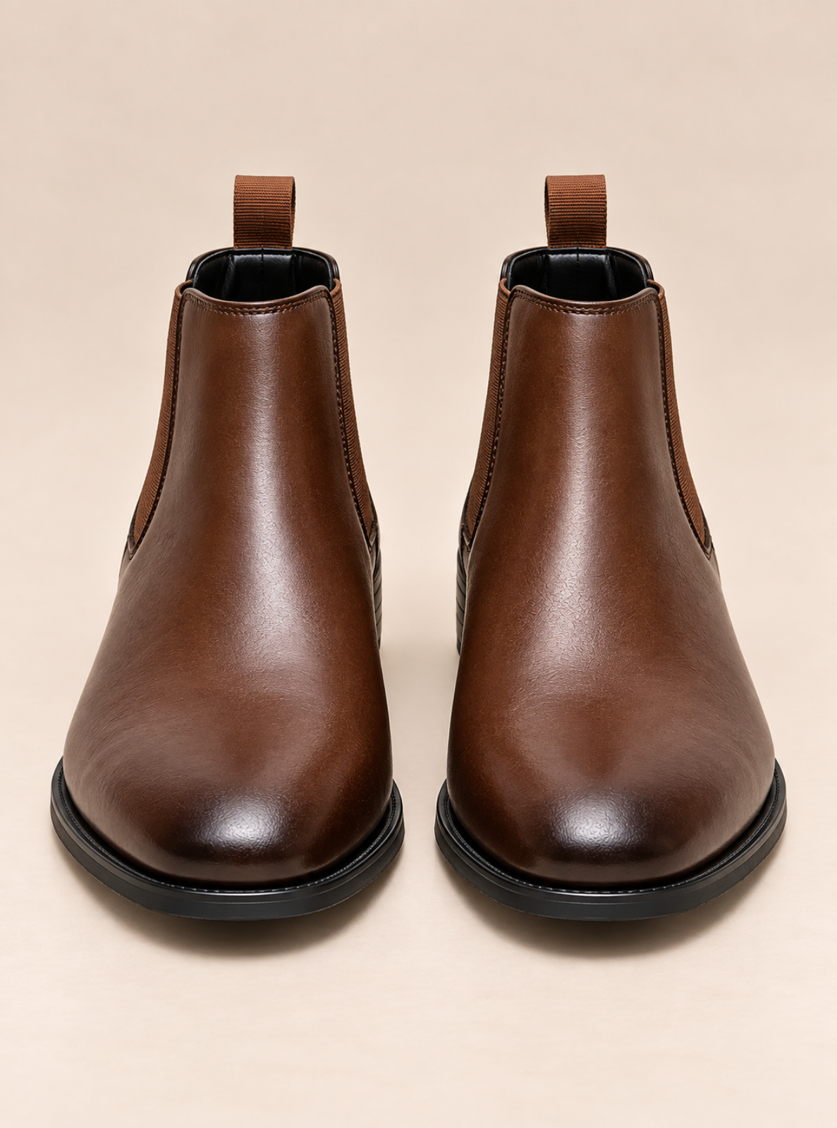 A pair of dark brown leather Chelsea boots with black elastic side panels and pull tabs, shown from a front-facing perspective on a light beige background.