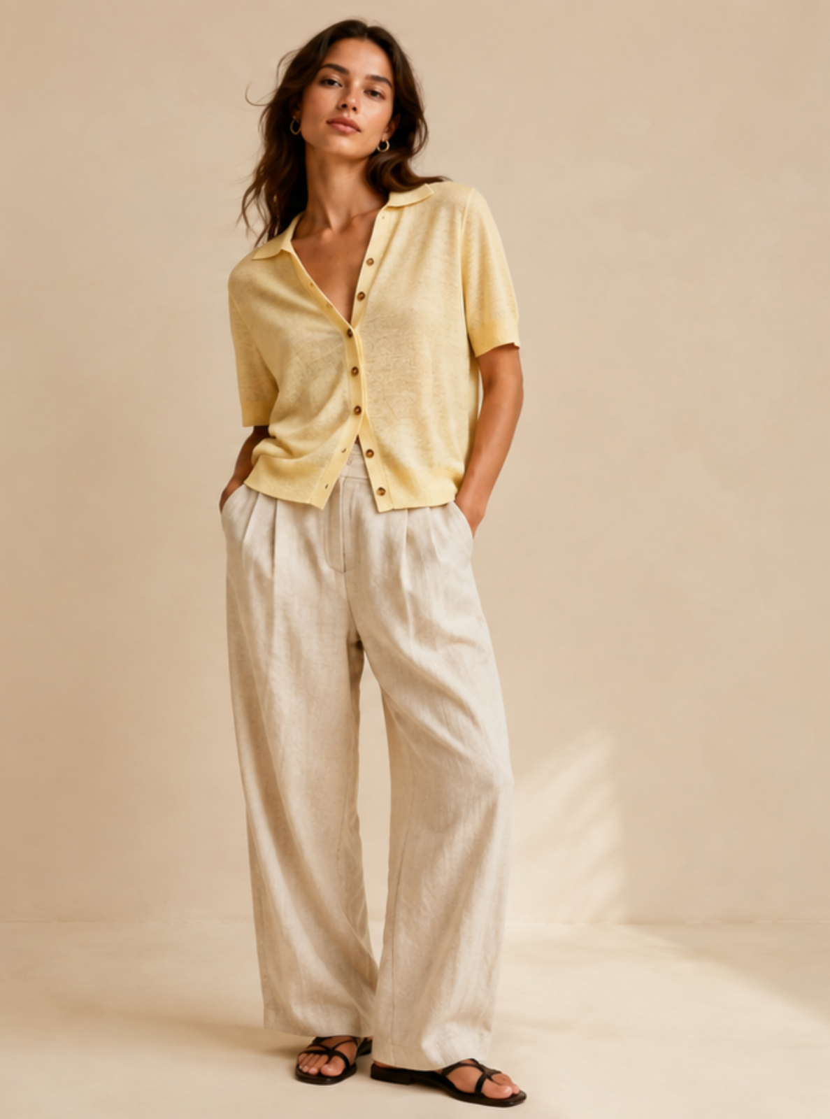 A woman modeling a pale yellow short-sleeve button-front top and off-white wide-leg pants against a warm beige studio backdrop.
