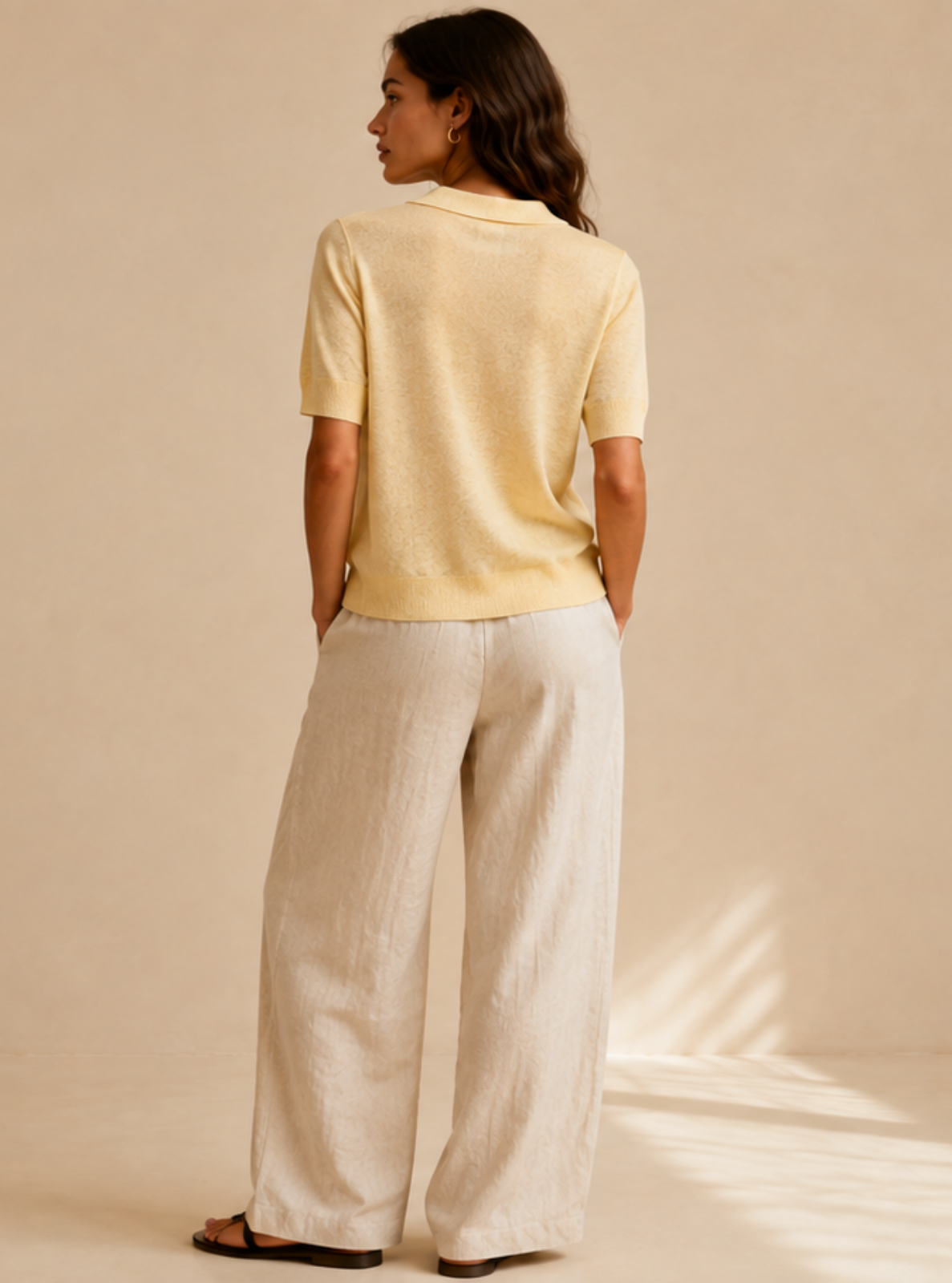 A woman stands with her back to the camera wearing a pale yellow short-sleeve top and light beige wide-leg trousers in a minimalist studio setting with warm beige walls and floor.