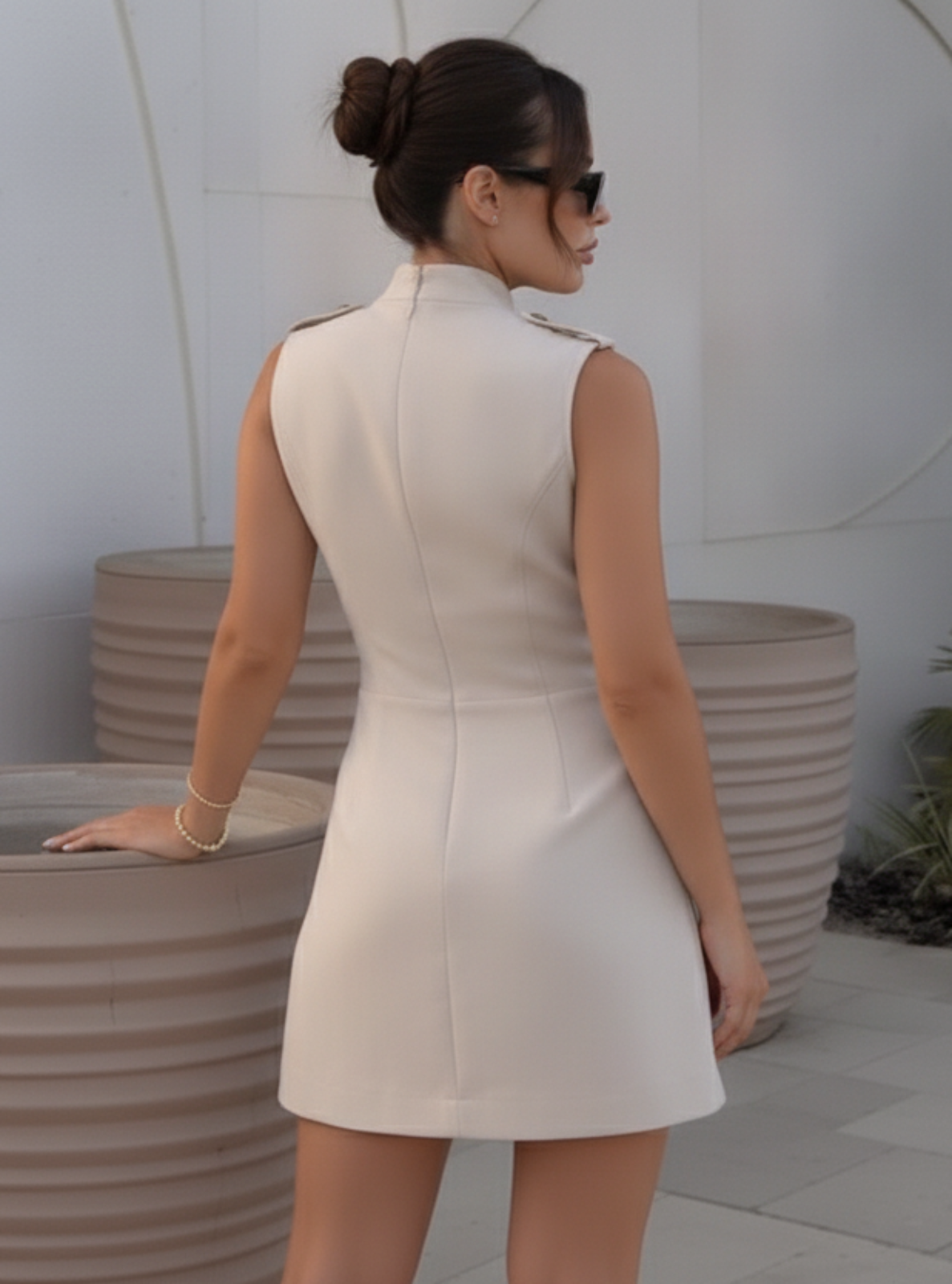 Woman in a white dress standing next to large planters against a white wall with circular patterns.