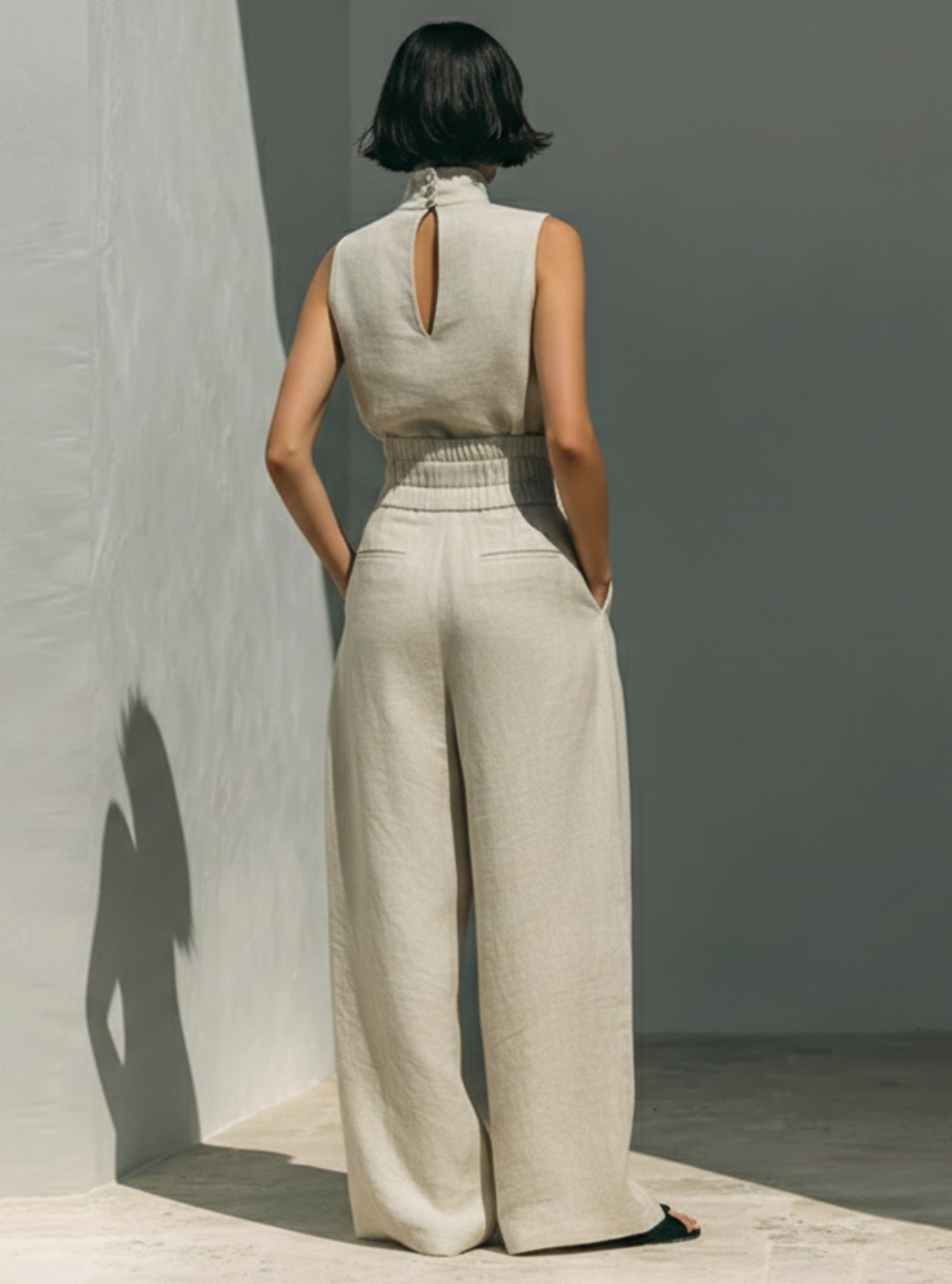 Woman wearing a sleeveless top and wide-leg pants standing against a plain wall.