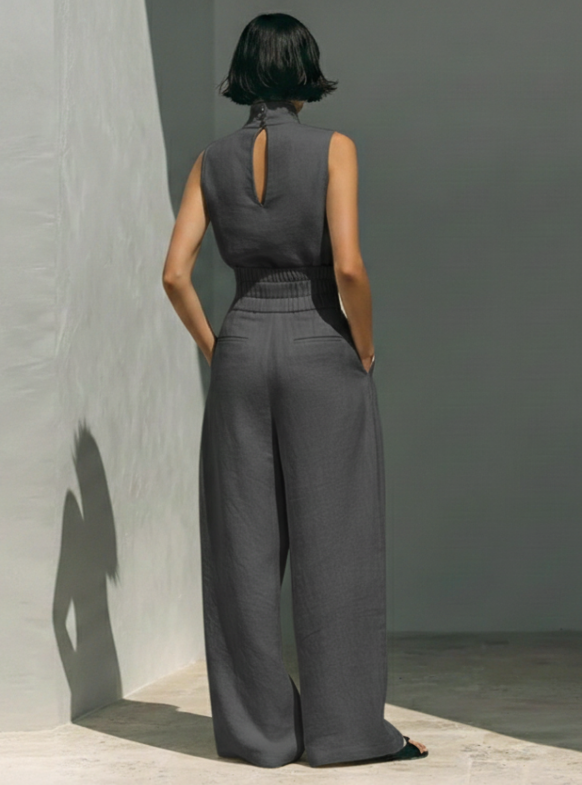 Person wearing a gray sleeveless jumpsuit standing against a plain wall.
