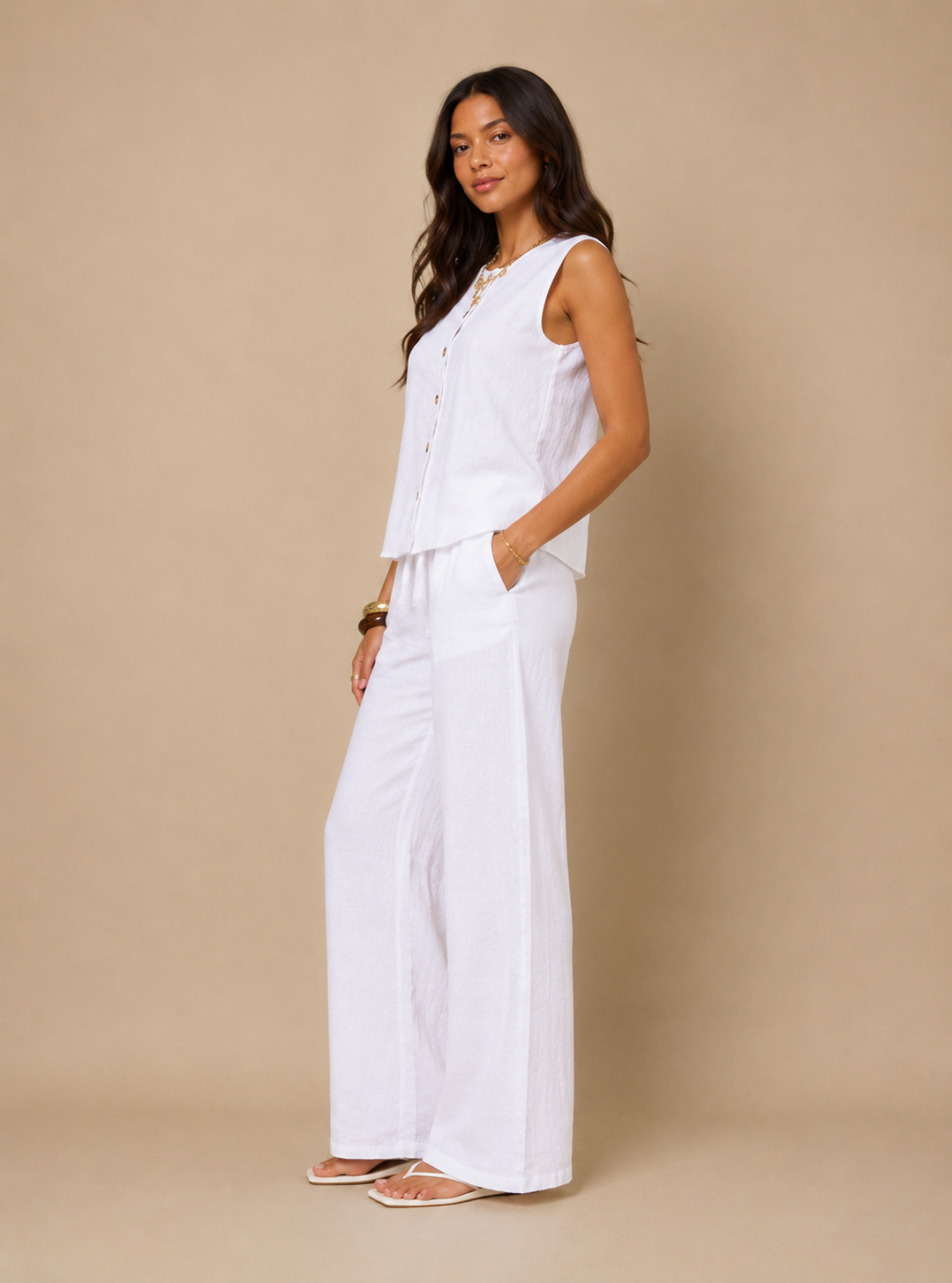 A woman modeling a white sleeveless button-front top and matching wide-leg pants set against a solid beige studio background.