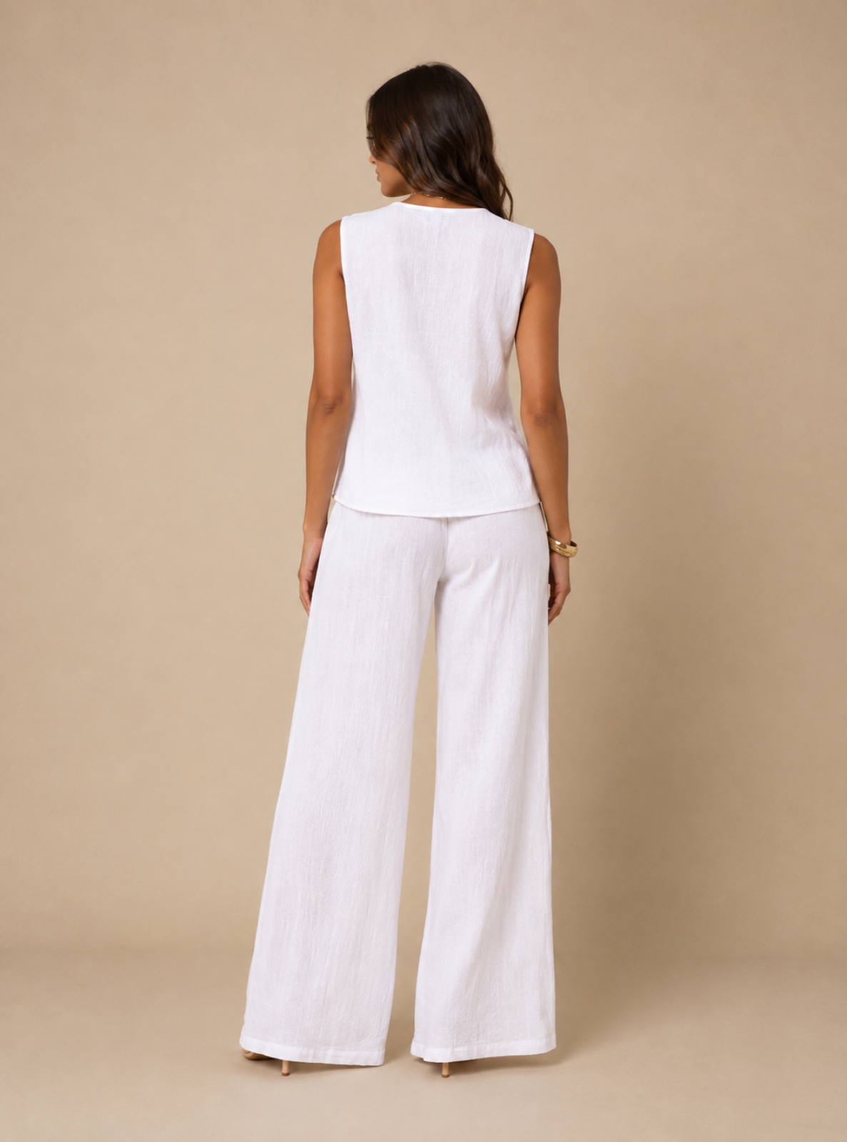 A woman viewed from the back wearing a sleeveless white top and matching wide-leg white pants against a plain beige studio backdrop.