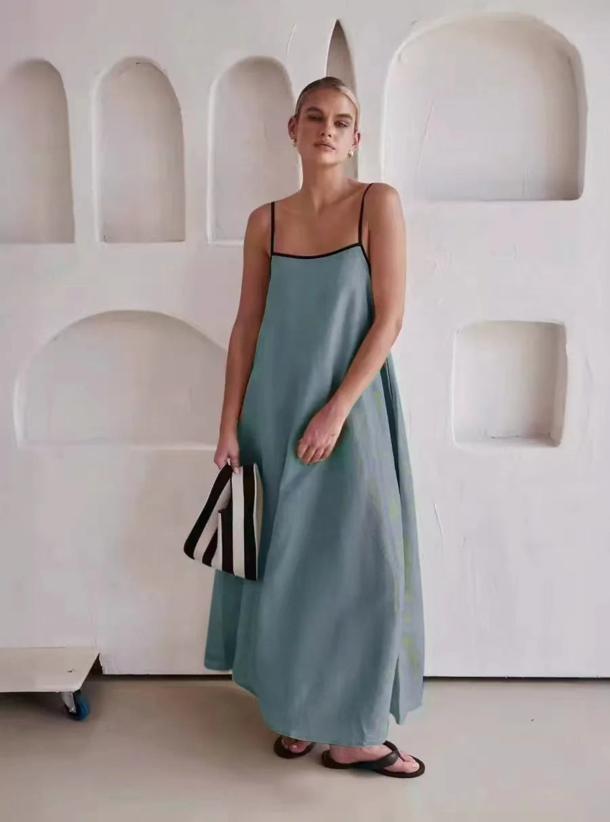 Woman in a long light blue dress holding a black and white striped bag against a minimalistic white wall.