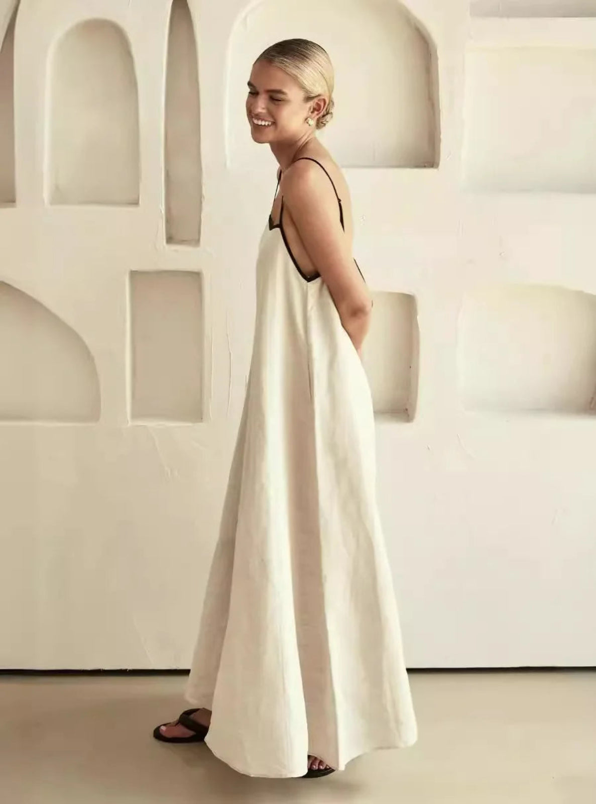 Woman in a long white dress standing in a minimalistic room with white walls and a white chair.